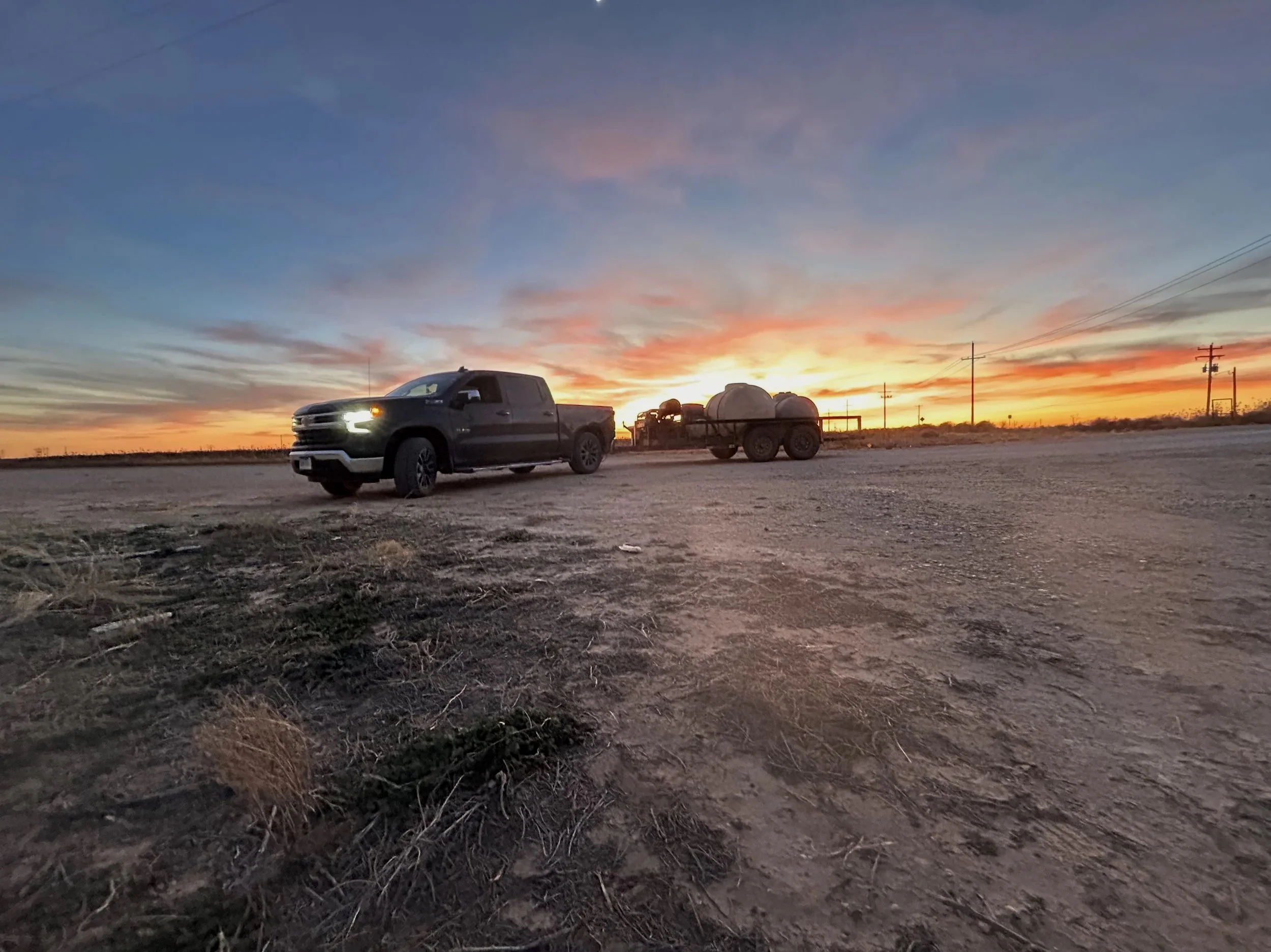 A black pickup truck pulling a trailer with tanks, parked on a flat dirt landscape during sunset, with power lines and a colorful sky in the background.
