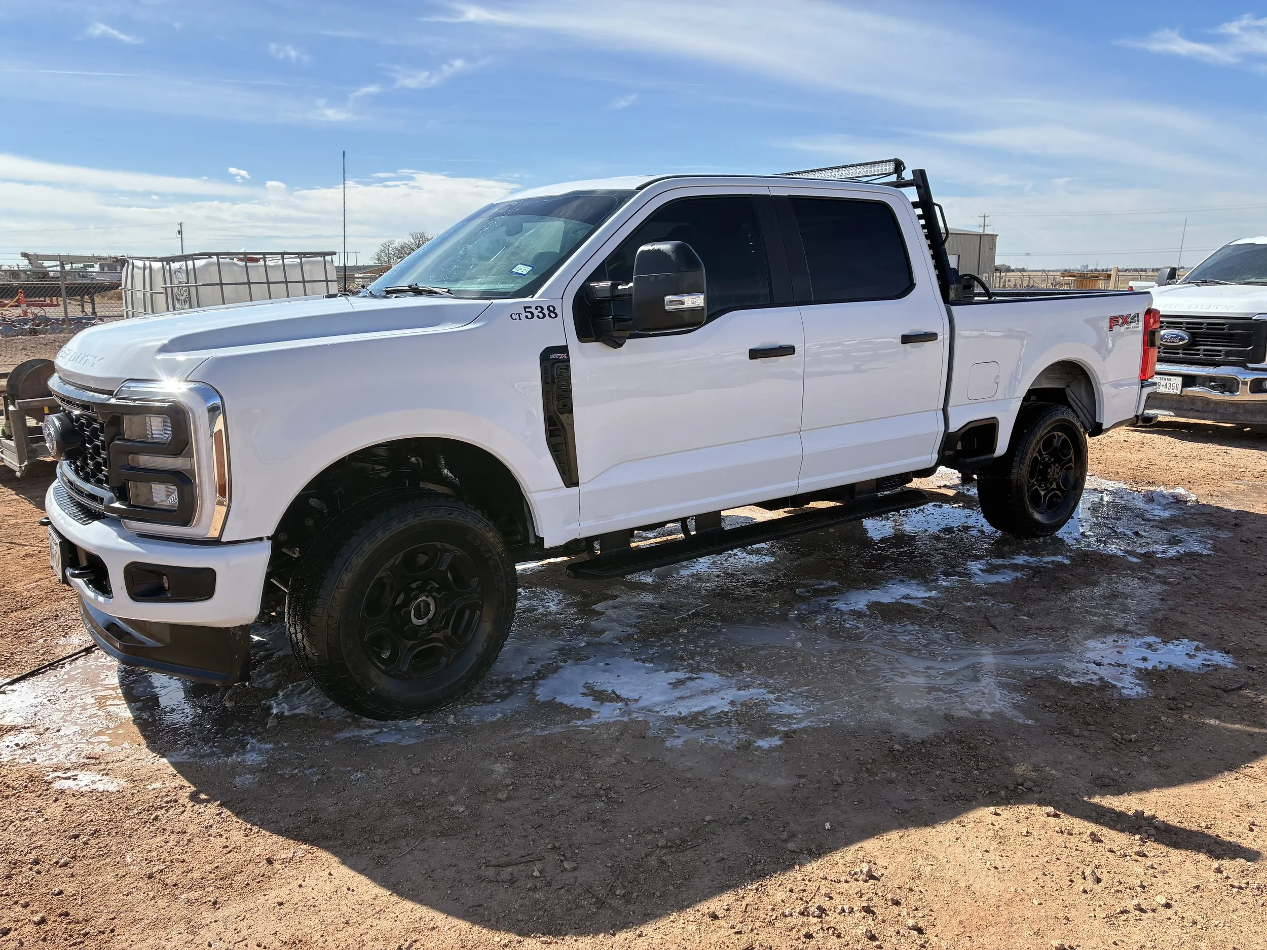 White pickup truck with black wheels parked on dirt ground with snow patches, under a blue sky with clouds, in a lot with other vehicles and fencing in the background.