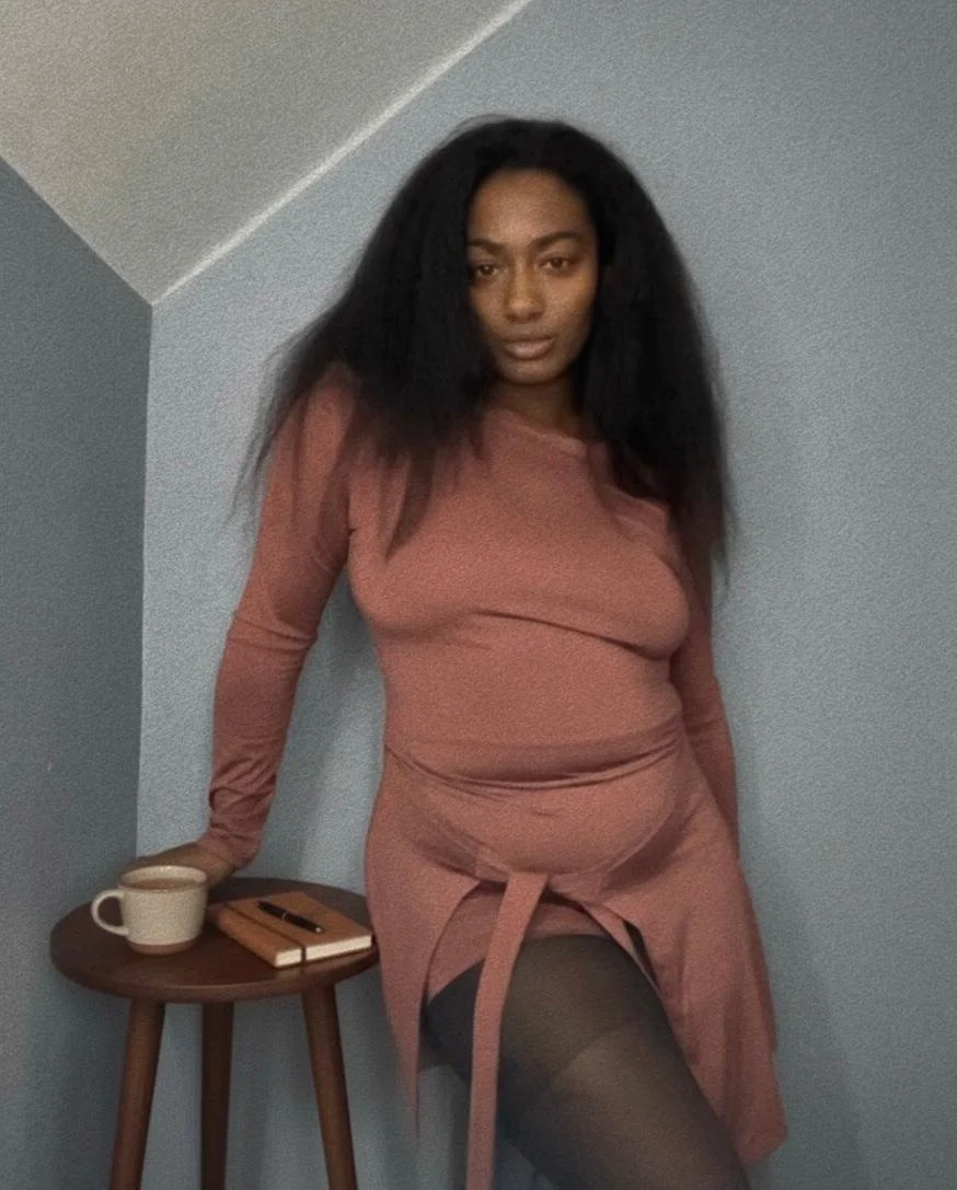 A woman with long, voluminous black hair leaning on a small round wooden table against a light blue wall, wearing a matching pink outfit and black sheer stockings, with a cup, notebook, and pen on the table.