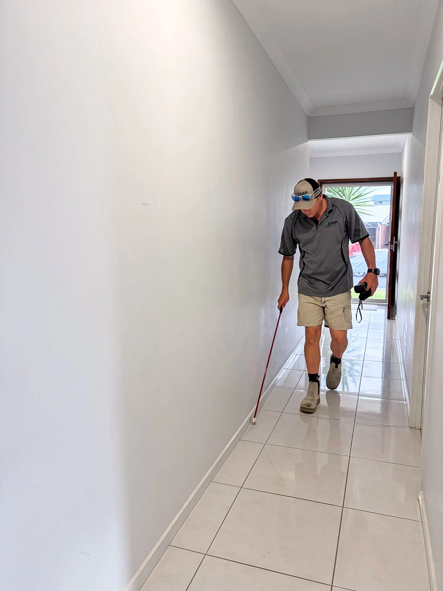 A man walking down a hallway while using a red laser level on the floor. He is wearing a gray polo shirt, beige shorts, boots, sunglasses, and a cap, and holds a camera or phone in his left hand.