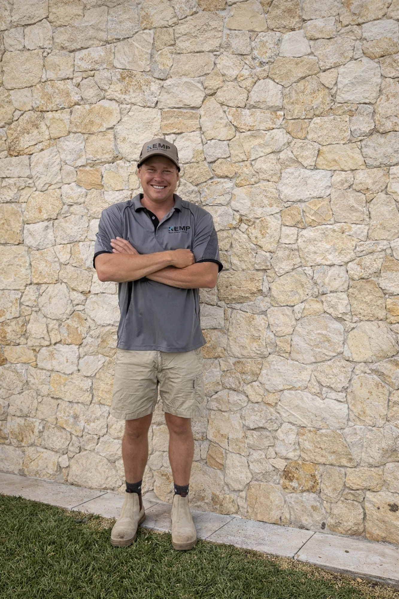 A man in work clothes standing with his arms crossed in front of a stone wall, smiling at the camera.