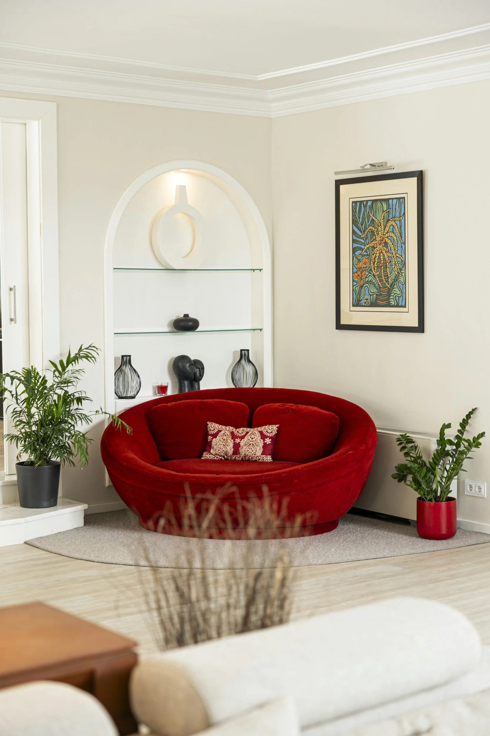 A living room with a red, rounded sofa with a decorative pillow, green potted plants on either side, a framed artwork on the wall, and built-in shelves with decorative vases and a sculpture.