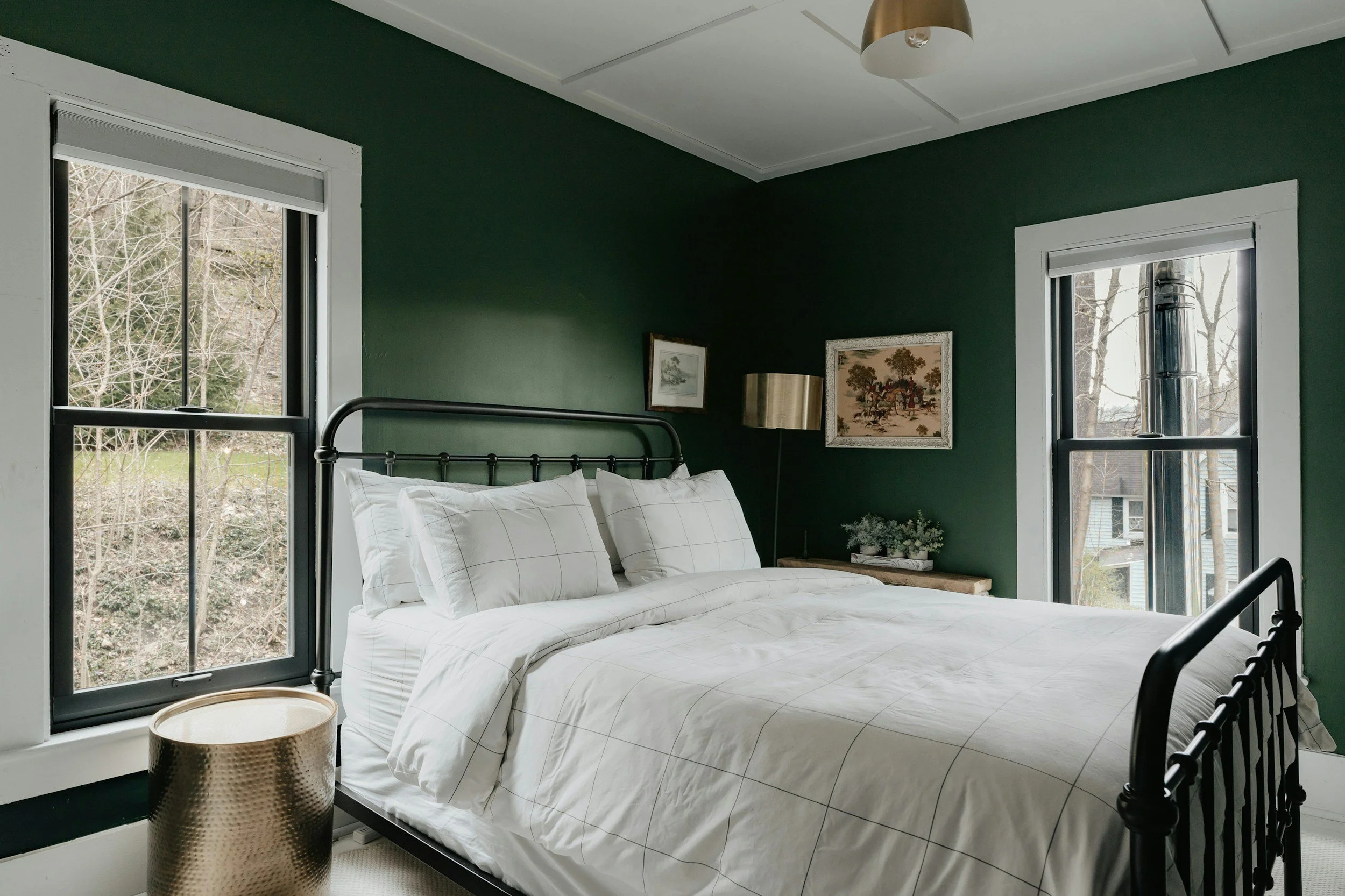 A bedroom with dark green walls, two windows with white trim, a black metal bed frame with white bedding and pillows, a gold bedside lamp, wall art above the bed, and a metallic textured side table near the bed.