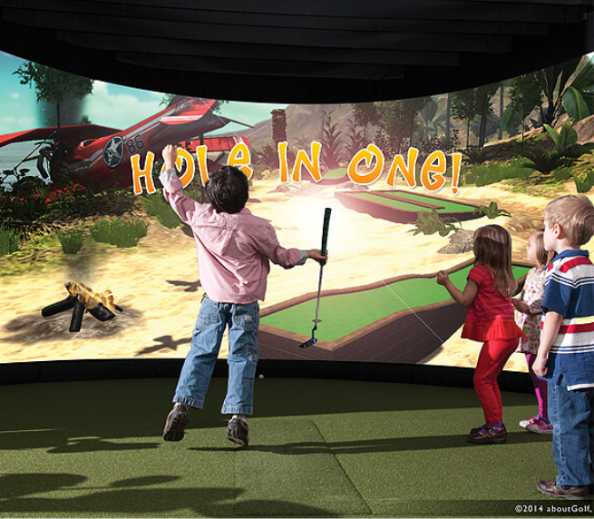 Children playing miniature golf inside a simulator with a desert-themed background that includes a helicopter and the text 'Hole in One!'