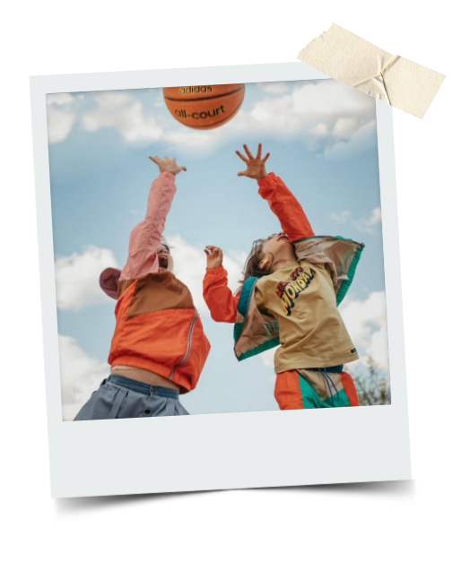 Two children playing basketball outdoors, reaching for the ball against a cloudy sky.