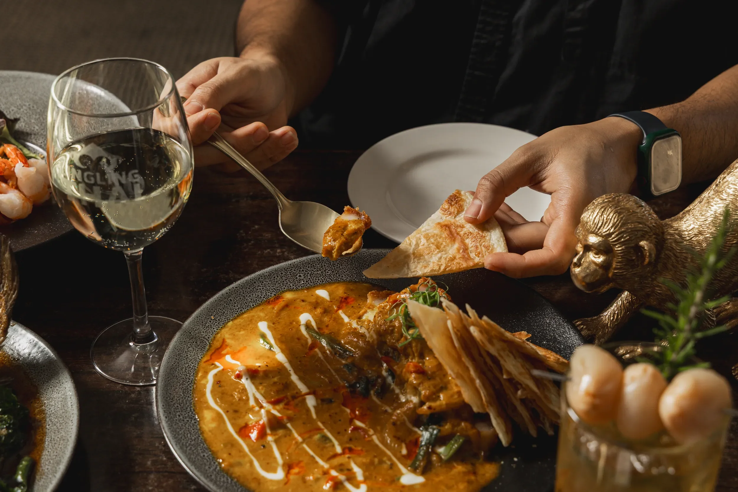 A person serving food from a black plate with curry onto a piece of naan bread in a dark dining setting, with a glass of white wine and another dish partially visible at the edge of the table.