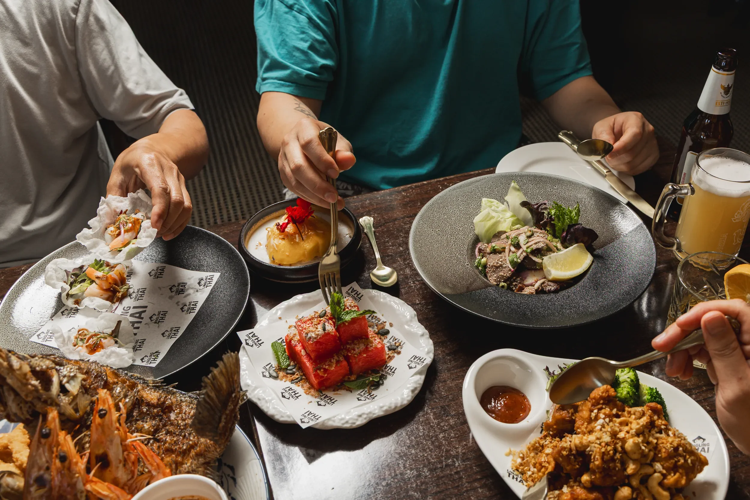 People eating various Asian dishes at a dining table, including seafood, barbecue, and side dishes, with drinks and condiments.