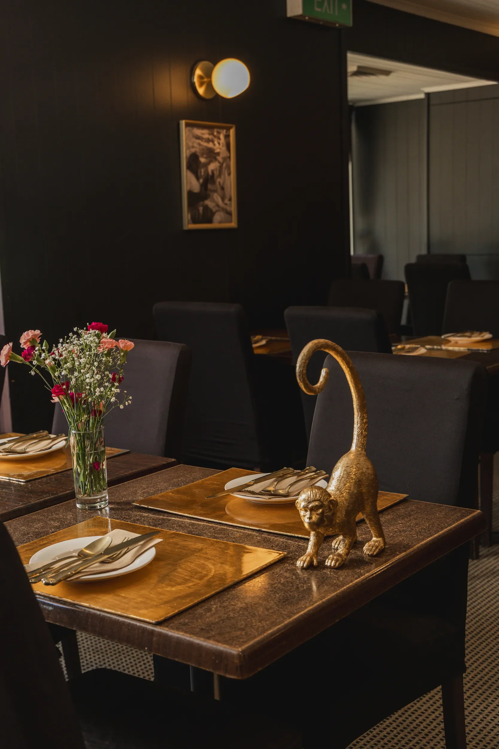 A dining table in a restaurant set with white plates, gold-colored cutlery, and gold placemats. A transparent vase with pink, red, and white flowers decorates the table. Gold-colored monkey figurine with a long curling tail is also on the table. The 