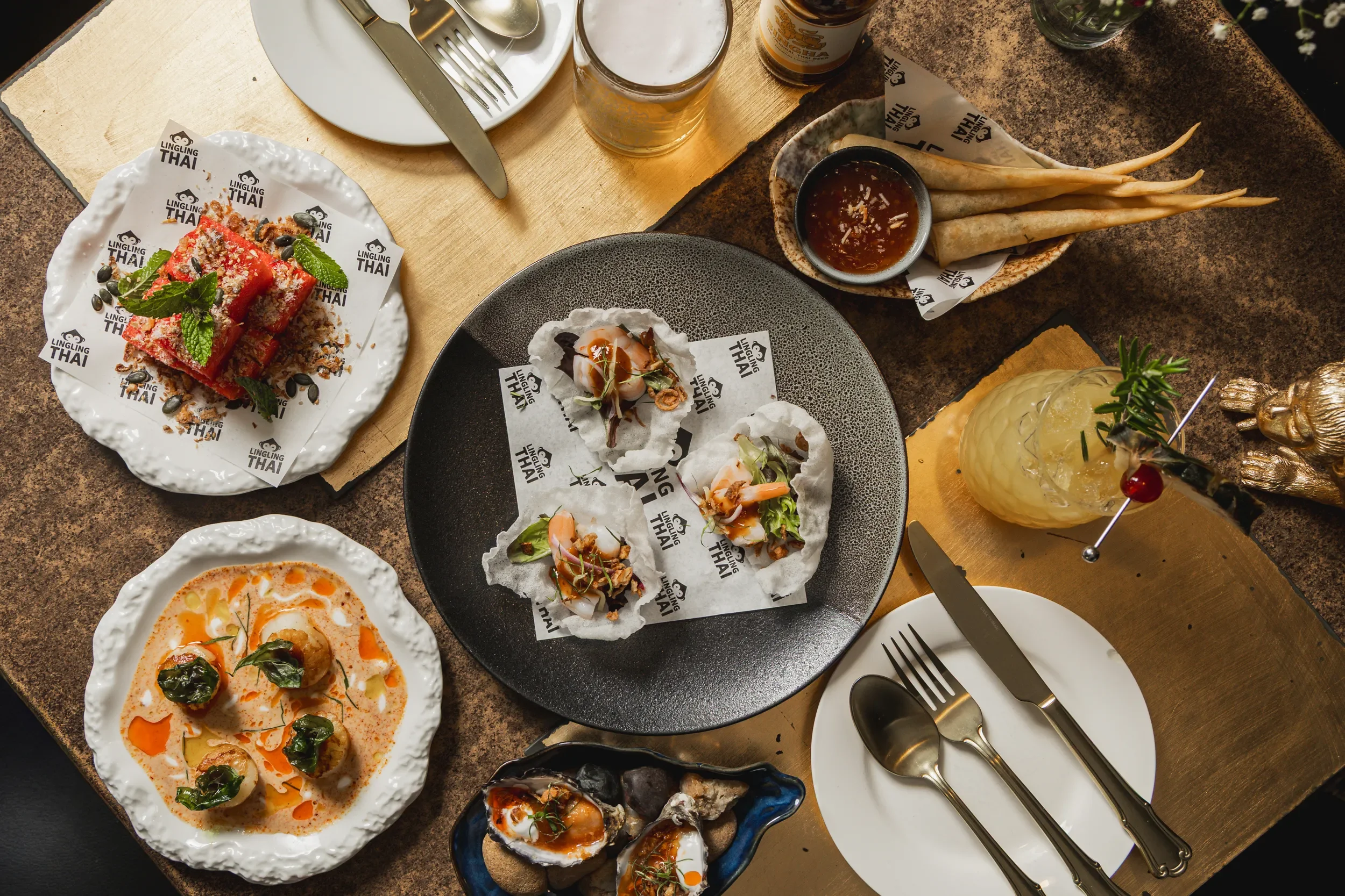 A table with various Thai dishes including sliced watermelon with mint, small seafood appetizers on parchment paper, a creamy dish with herbs, and a plate of oysters, along with a cocktail, beer, and dipping sauces, set with plates, cutlery, and a wo