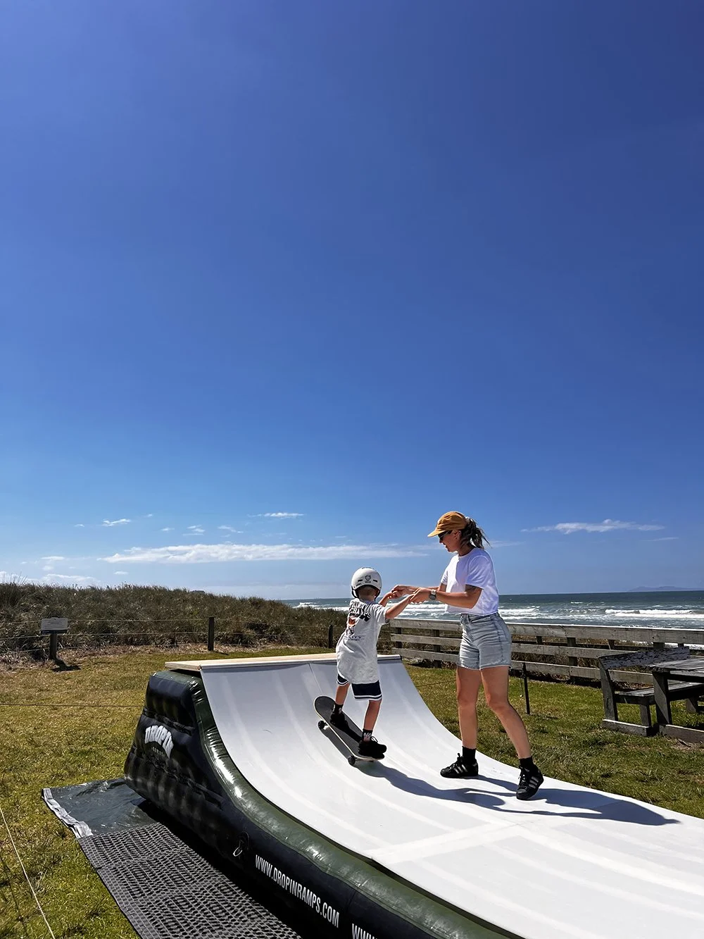 A woman helping a young boy learn to skateboard on a mini ramp outdoors near the beach on a sunny day.