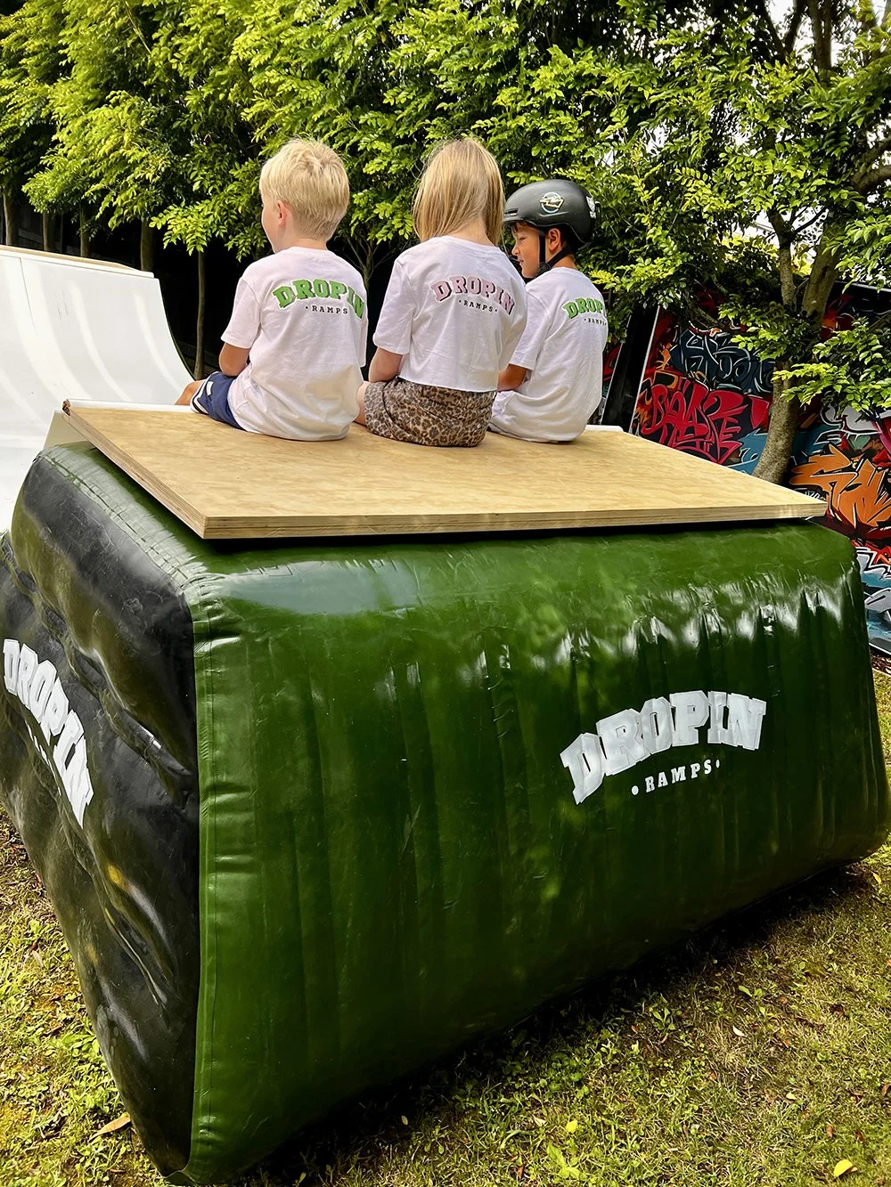 Three children sitting on a wooden platform with their backs facing the camera, wearing white t-shirts with 'DROPLIN RAMPS' printed on them, in an outdoor skate park with trees and graffiti in the background.