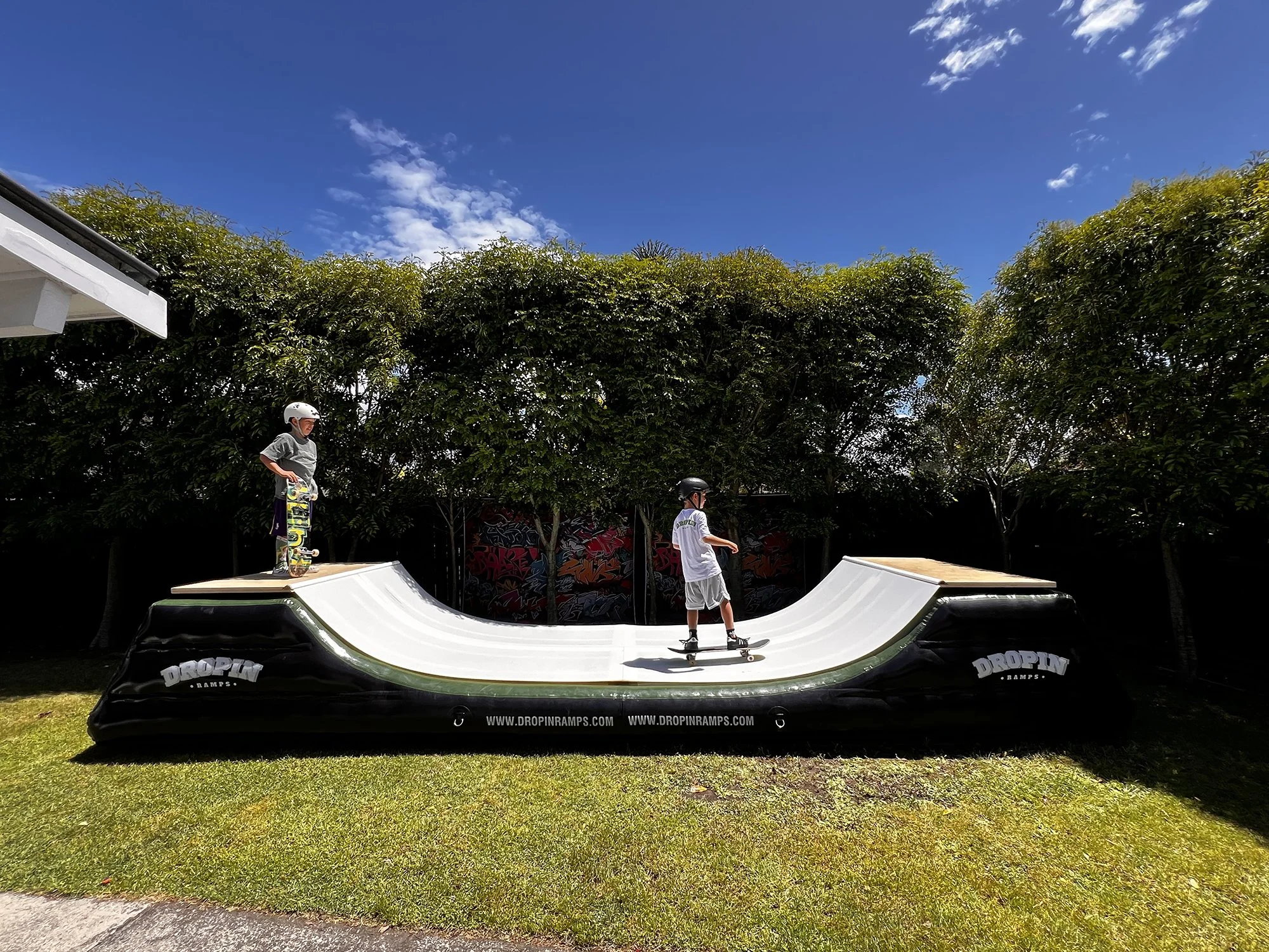 Two children skateboarding on a mini ramp outdoors under a blue sky with some clouds, surrounded by green trees.