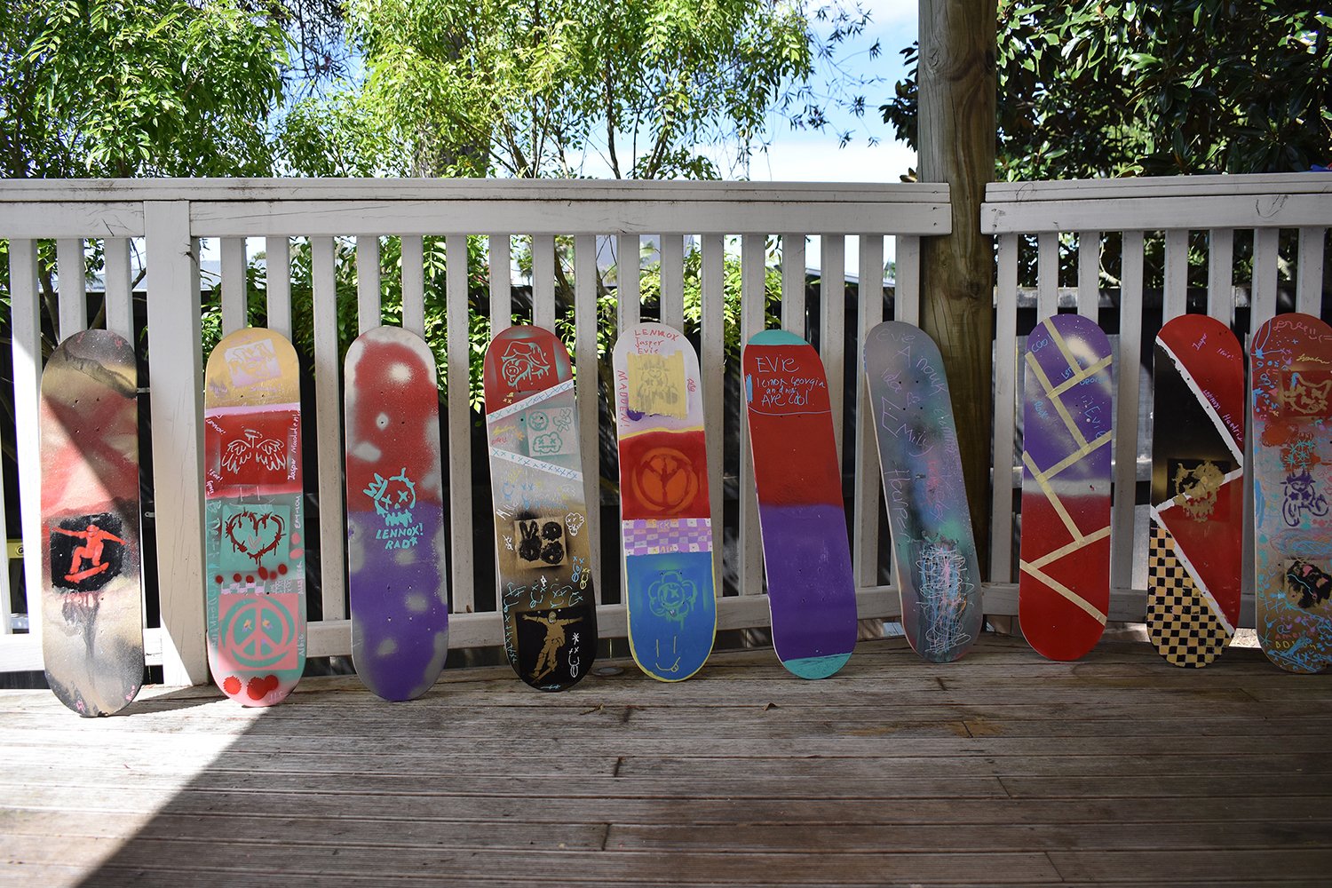A row of skateboards with colorful designs and each covered in graffiti, leaning against a white fence on a wooden deck. Green trees and blue sky are visible in the background.