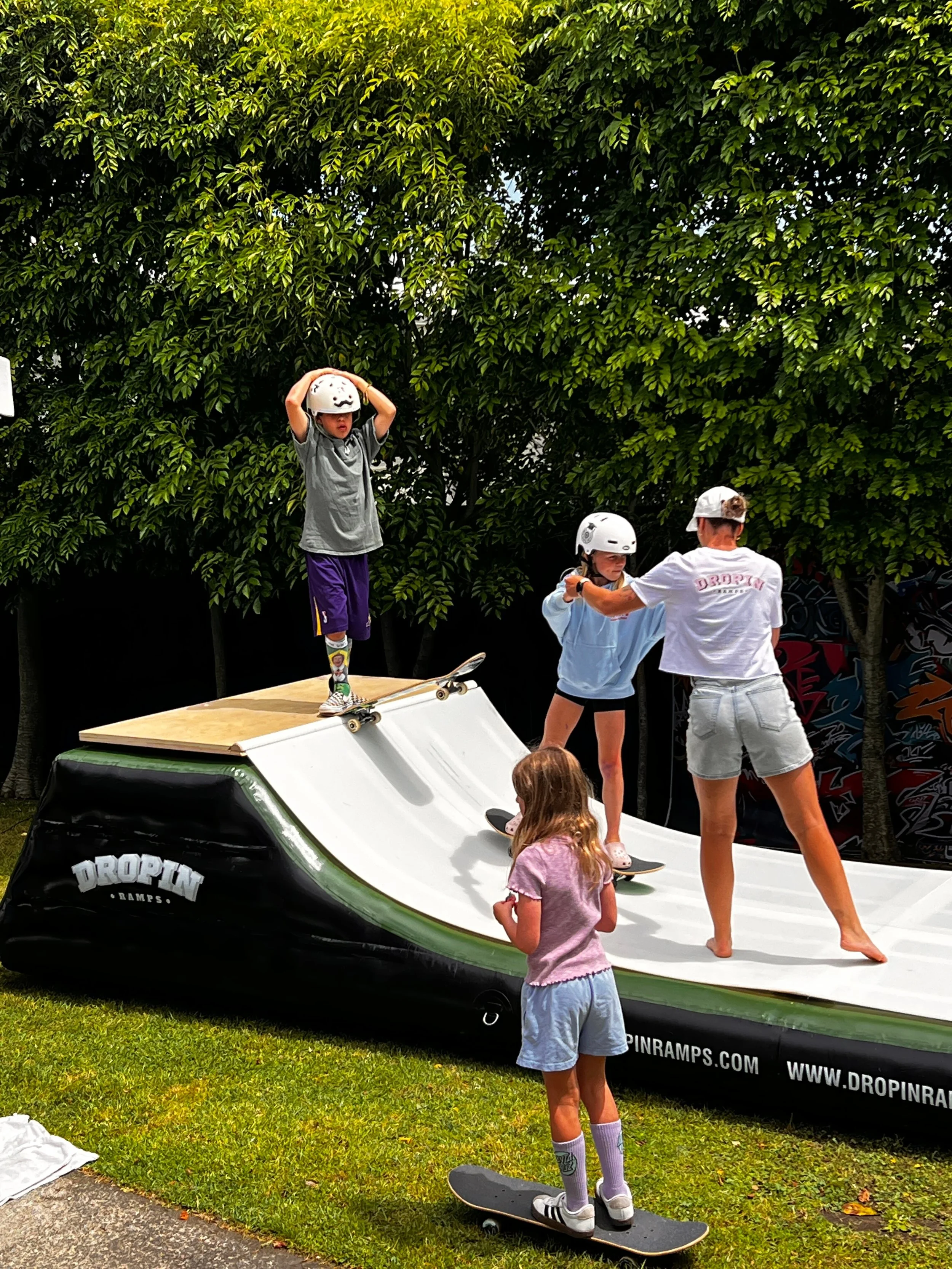 Kids learning skateboarding on an inflatable ramp, with an instructor assisting, outdoors with green trees in the background.