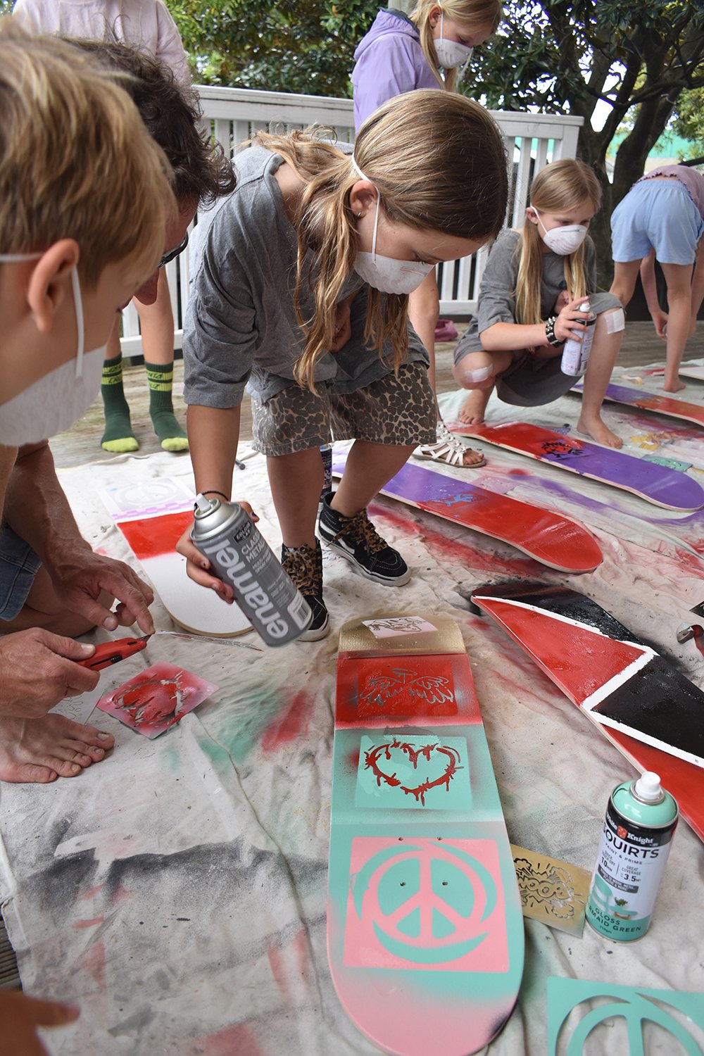 Children and adults wearing masks spray-painting and decorating skateboards outdoors on a large cloth-covered surface.