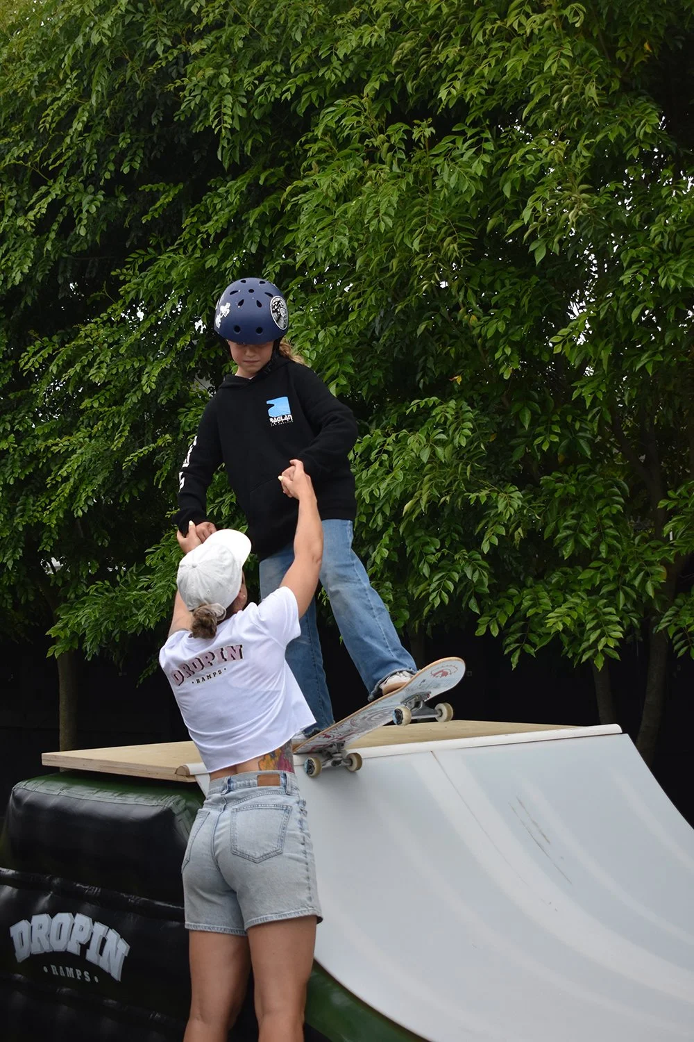 A girl wearing a white cap, a white t-shirt with the word 'DROPIN' on the back, and gray shorts is helping a boy wearing a black hoodie, blue jeans, and a helmet while standing on a skateboard on a ramp.