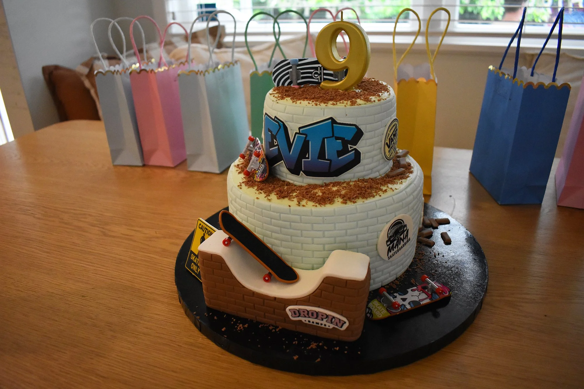 A two-tiered deviantart-themed birthday cake with skateboards and graffiti decorations, topped with a gold number 9 candle, surrounded by colorful gift bags, on a wooden table.