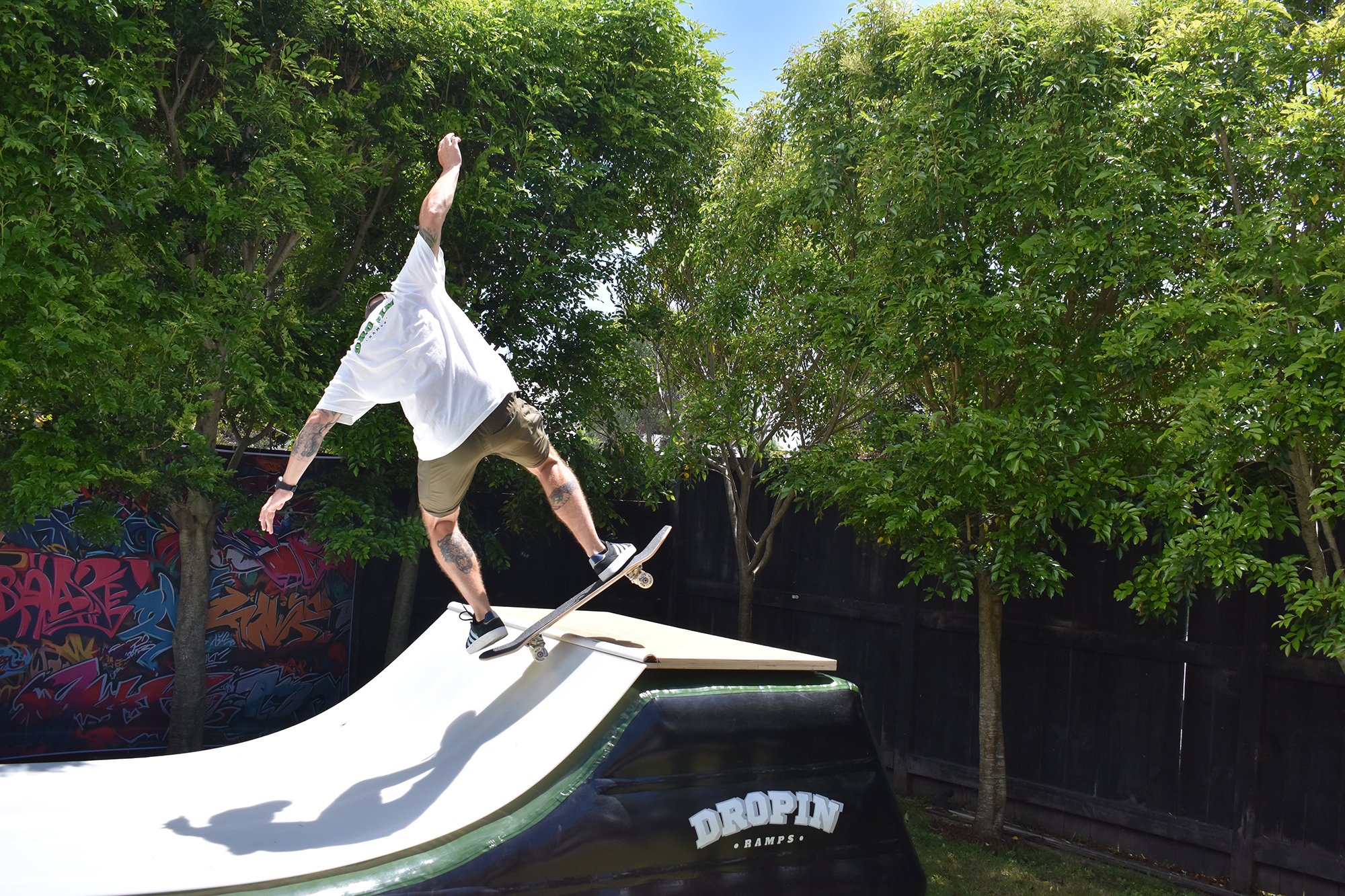 A man with tattoos on his arms and legs is skateboarding on a ramp outdoors, surrounded by trees and a wooden fence. He is wearing a white T-shirt, shorts, and sneakers, performing a trick on the skateboard.