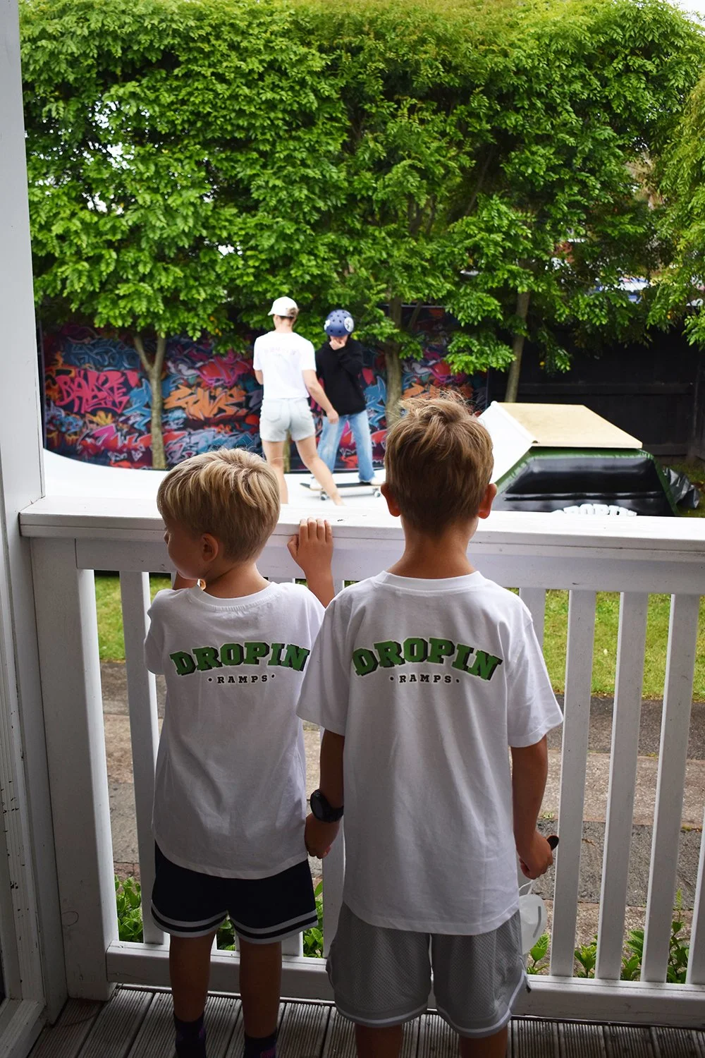 Two young boys stand on a porch, watching two older boys riding scooters and skateboarding in a backyard. The backyard has green trees and a graffiti-covered wall in the background.