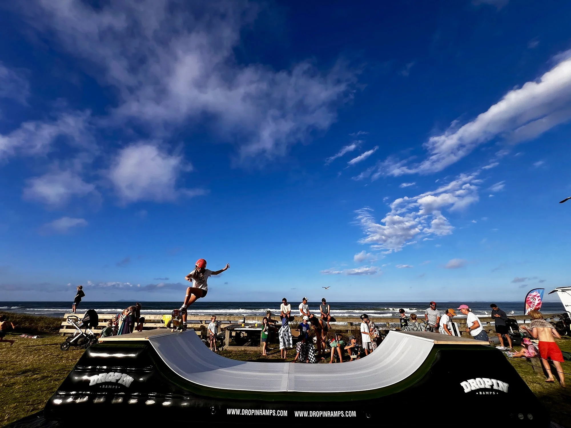 Children and adults at the beach enjoying a skateboarding ramp on a sunny day with blue sky and few clouds.