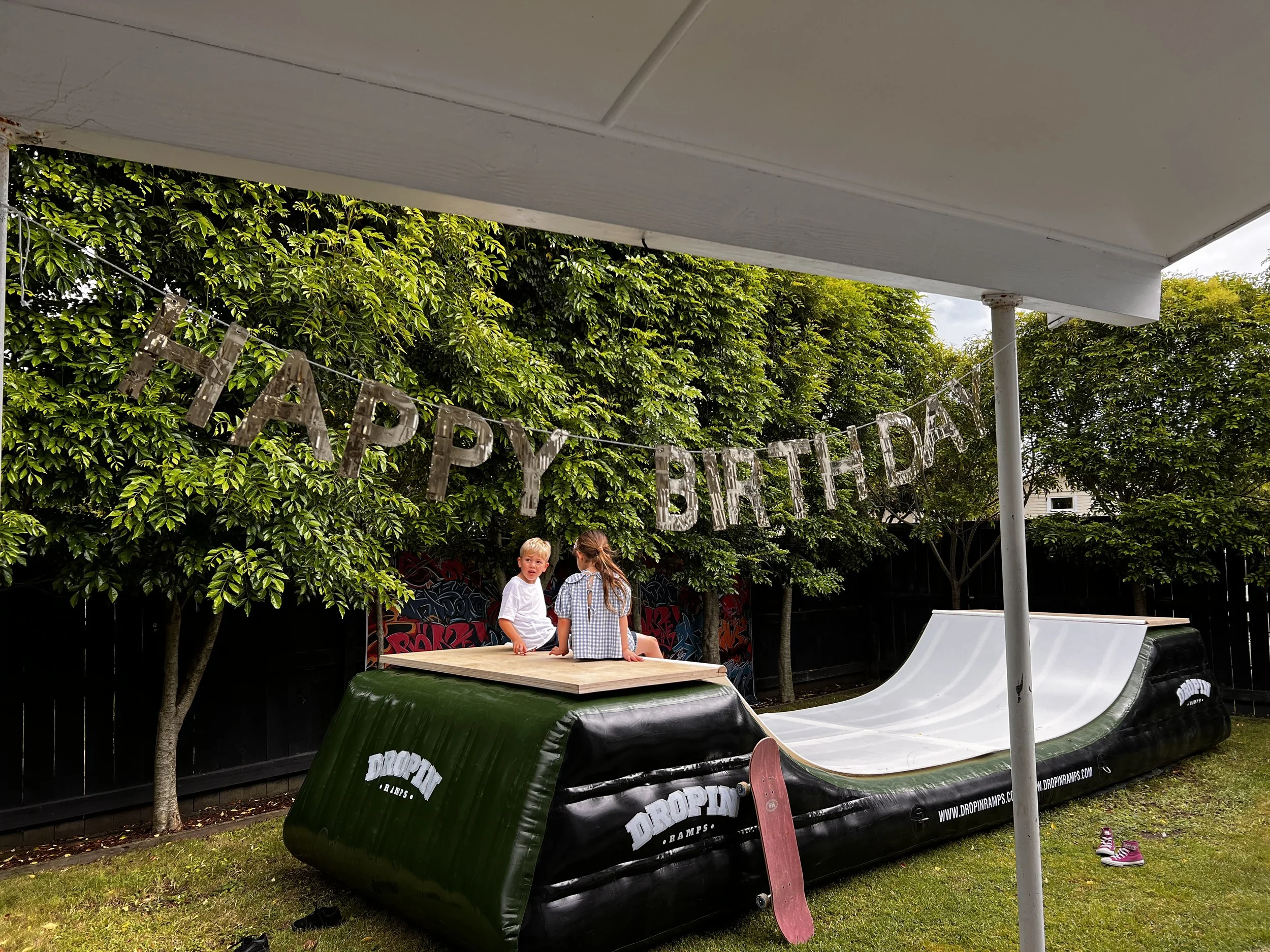 Two children playing on an inflatable skateboard ramp in a backyard, with a
