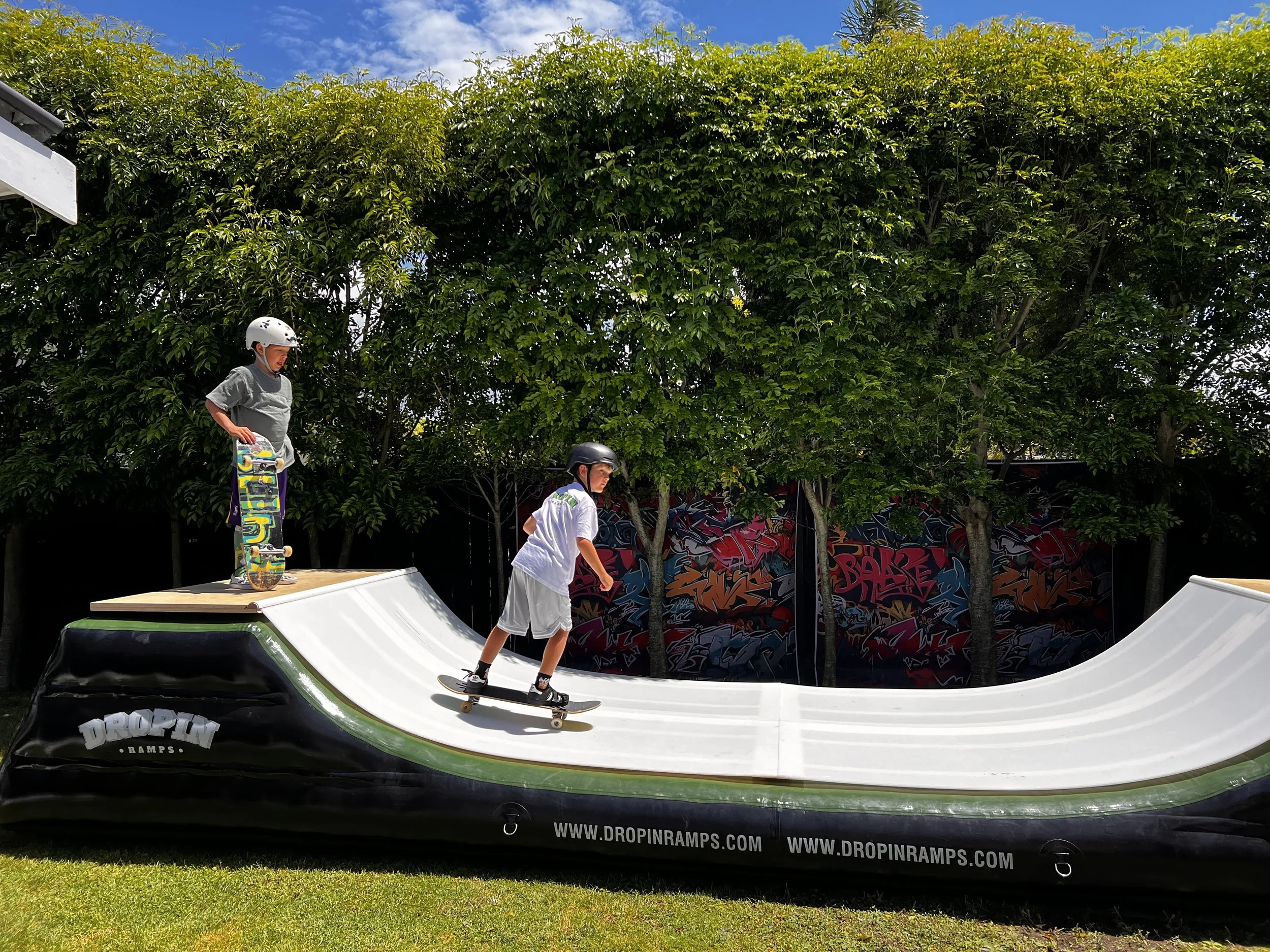 Two children wearing helmets skateboarding on a ramp with a green and black exterior, under a partly cloudy sky with trees in the background.