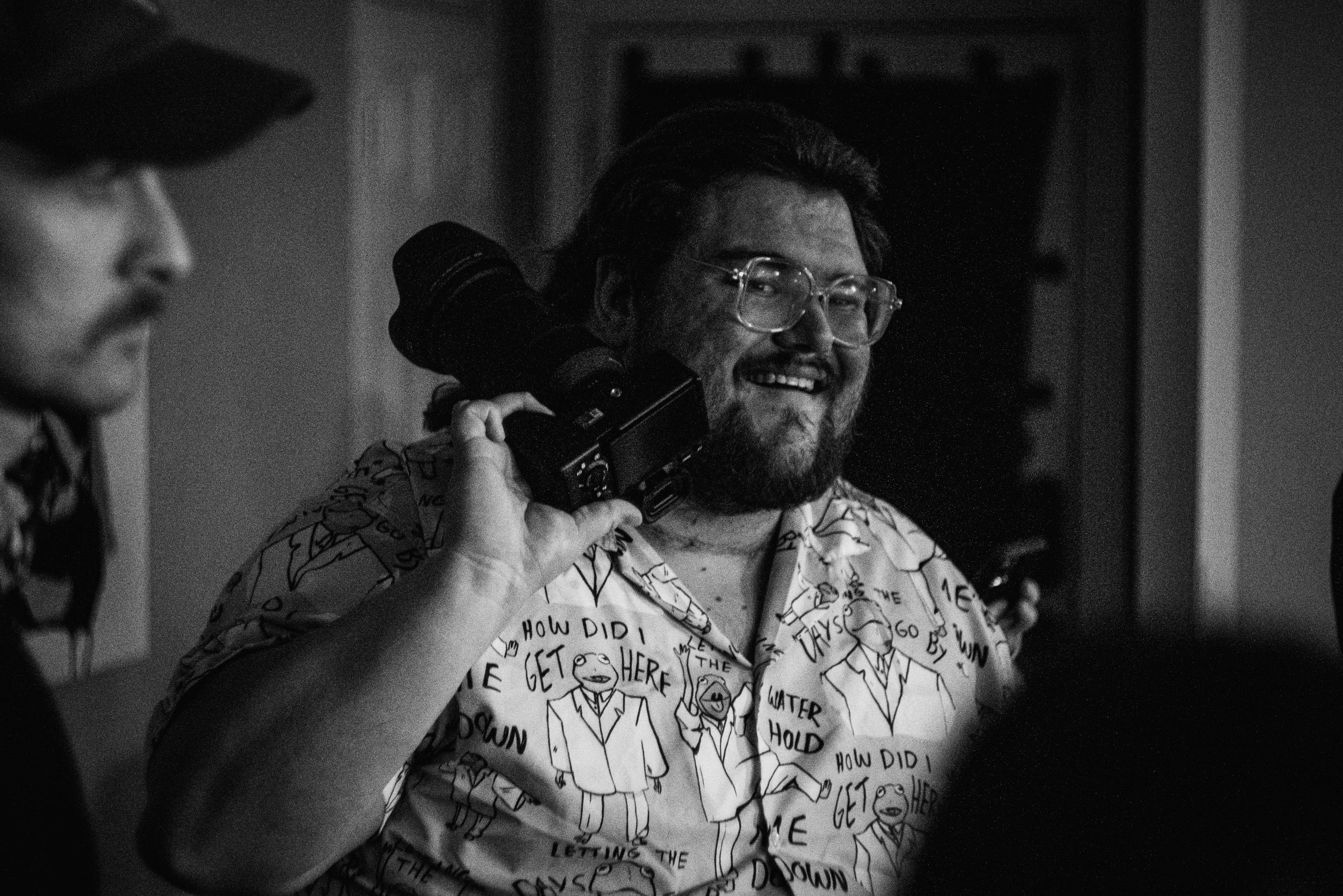 A man with glasses, a beard, and a patterned shirt is smiling while holding a camera on his shoulder.