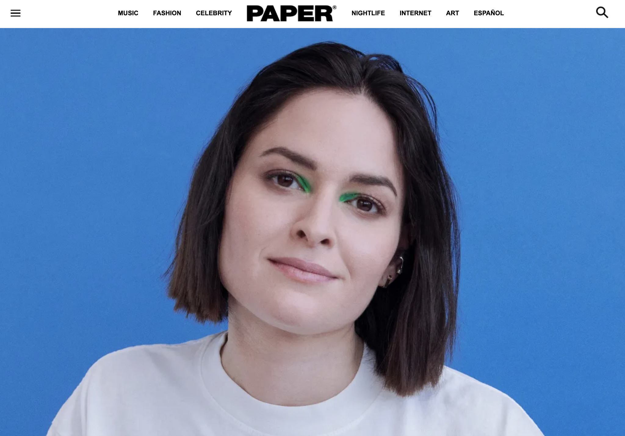 A young woman with short dark hair and light skin, smiling with green eyeliner, wearing a white t-shirt against a solid blue background.