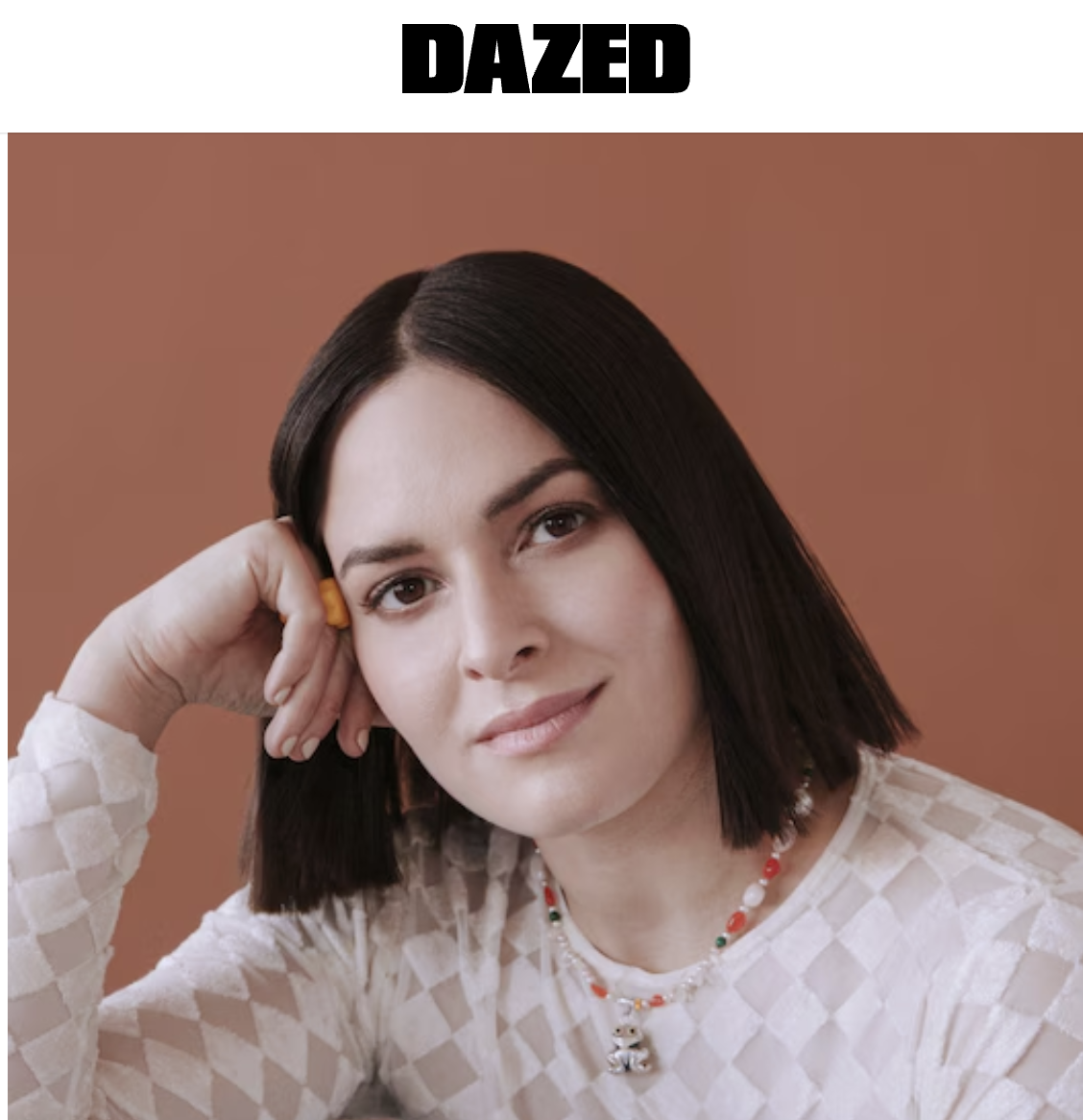 A woman with short dark hair and light skin, wearing a white checkered top, a colorful beaded necklace, and resting her head on her hand, against a brown background.