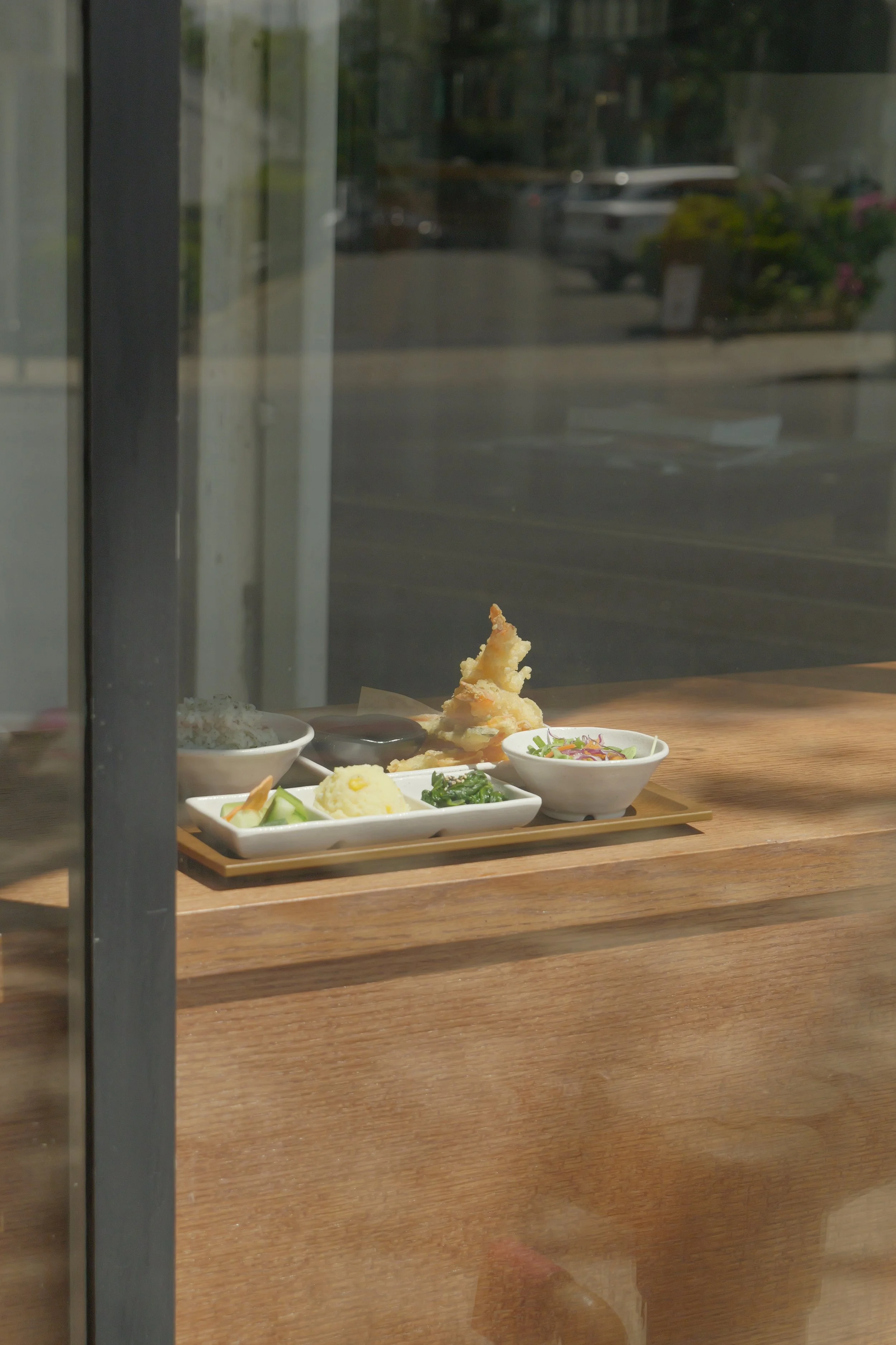A tray of Japanese-style lunch set dishes including sushi, tempura, rice, and vegetables seen through a restaurant window.