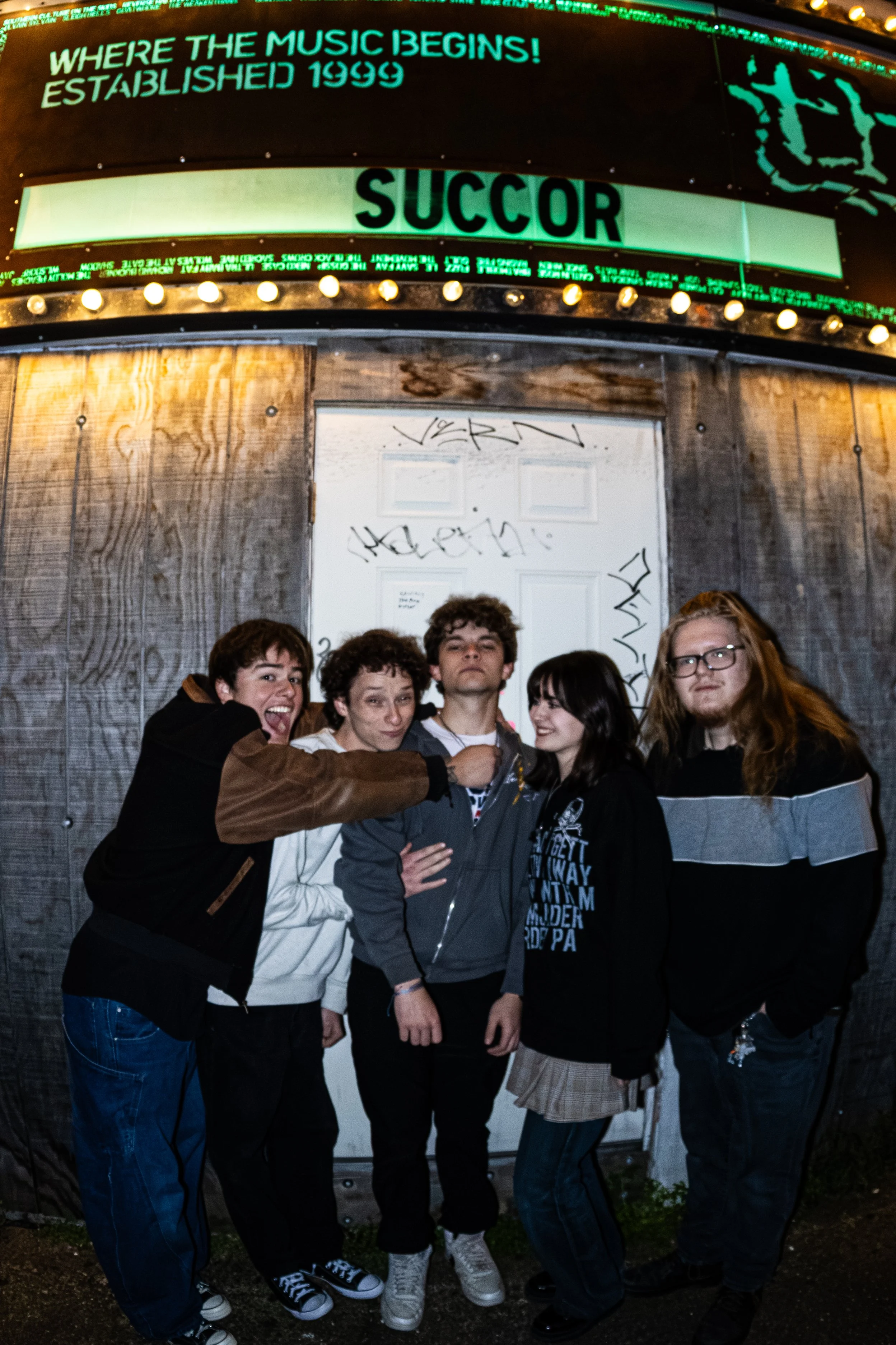 A group of five young people posing in front of a graffiti-covered door and an illuminated marquee advertising Succor at the venue The End, established in 1999.