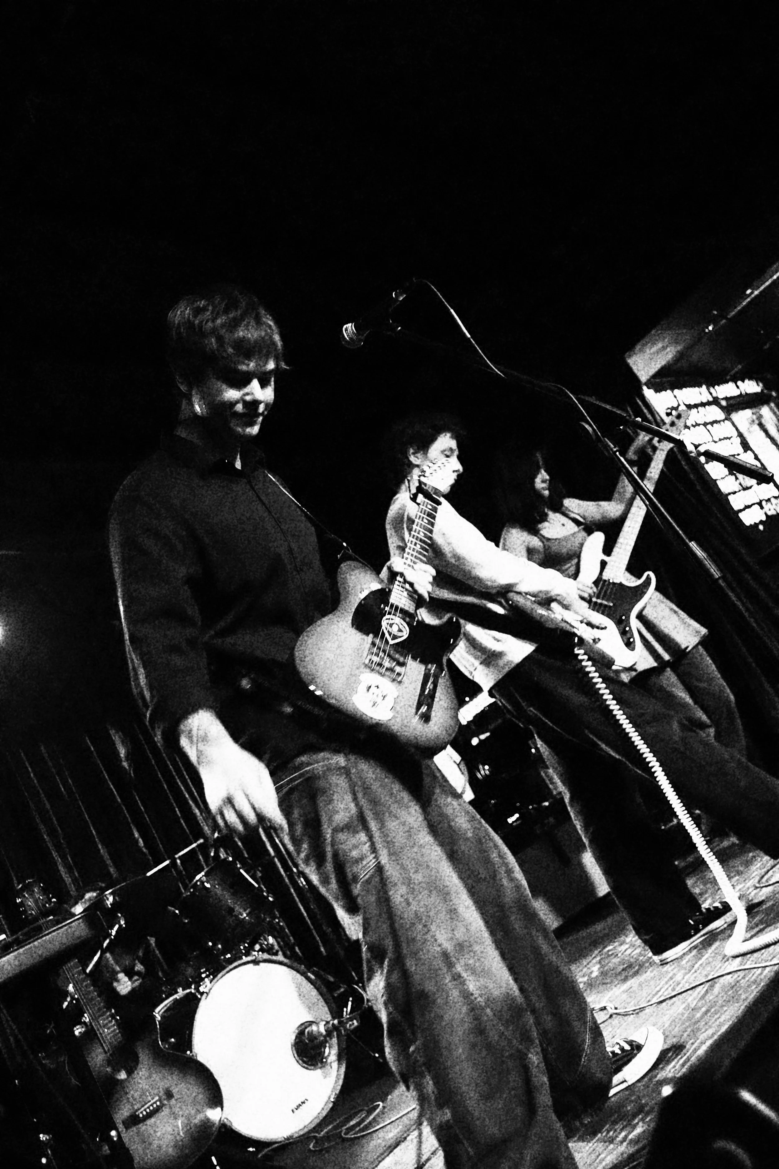 Black and white photo of a live band performing on stage, featuring three musicians on guitars and a man on drums, in a dimly lit venue.