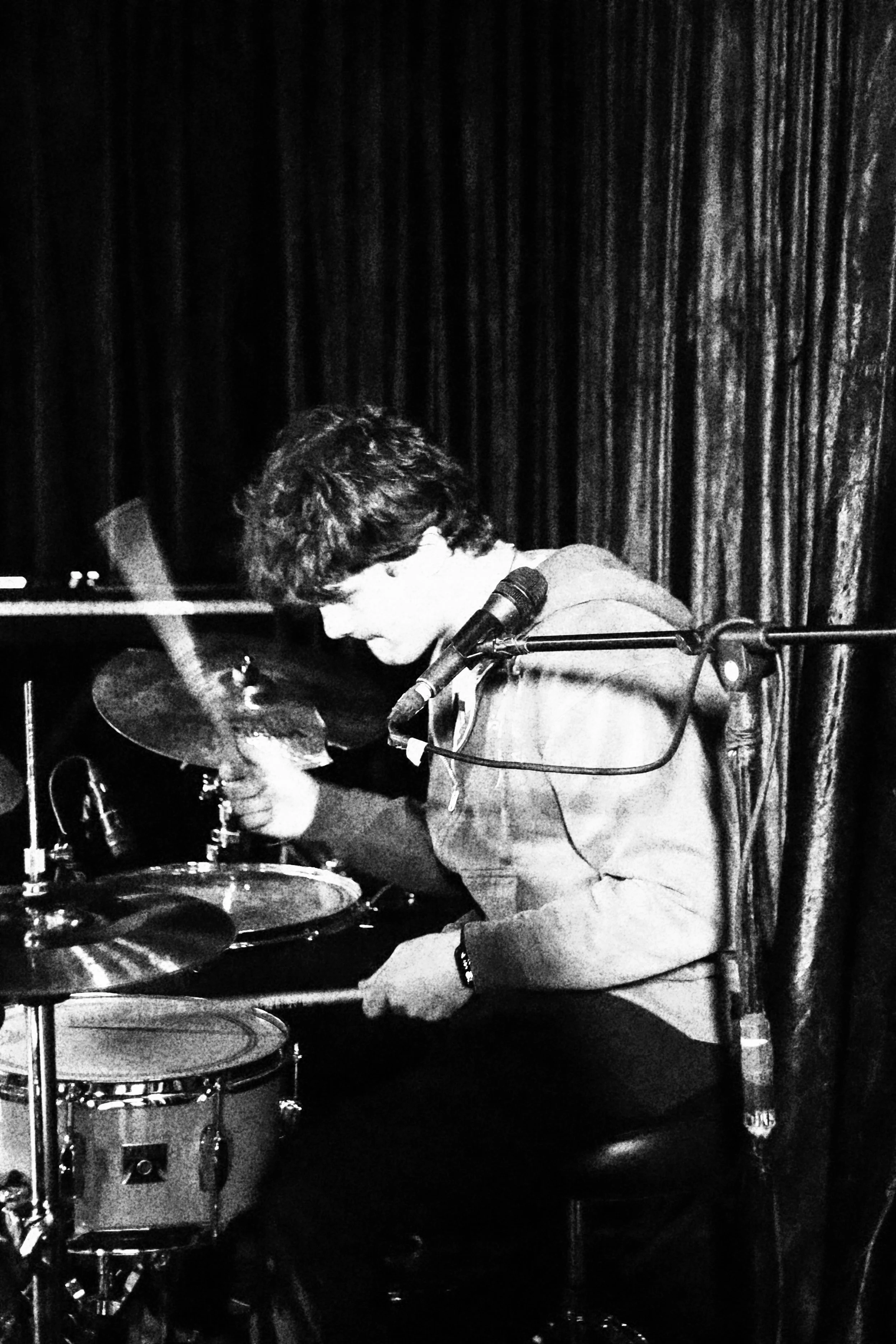A young man with dark hair playing drums on stage, behind a microphone, with dark curtains in the background.