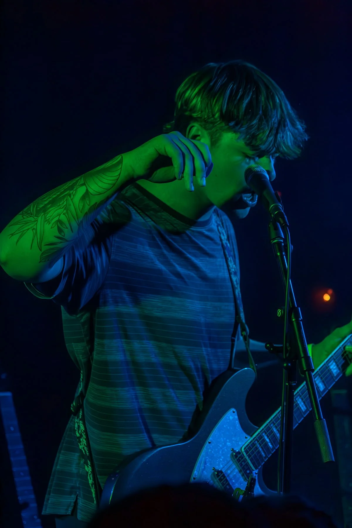 A young man with tattoos on his arm is singing into a microphone while playing an electric guitar on stage under green and blue stage lighting.