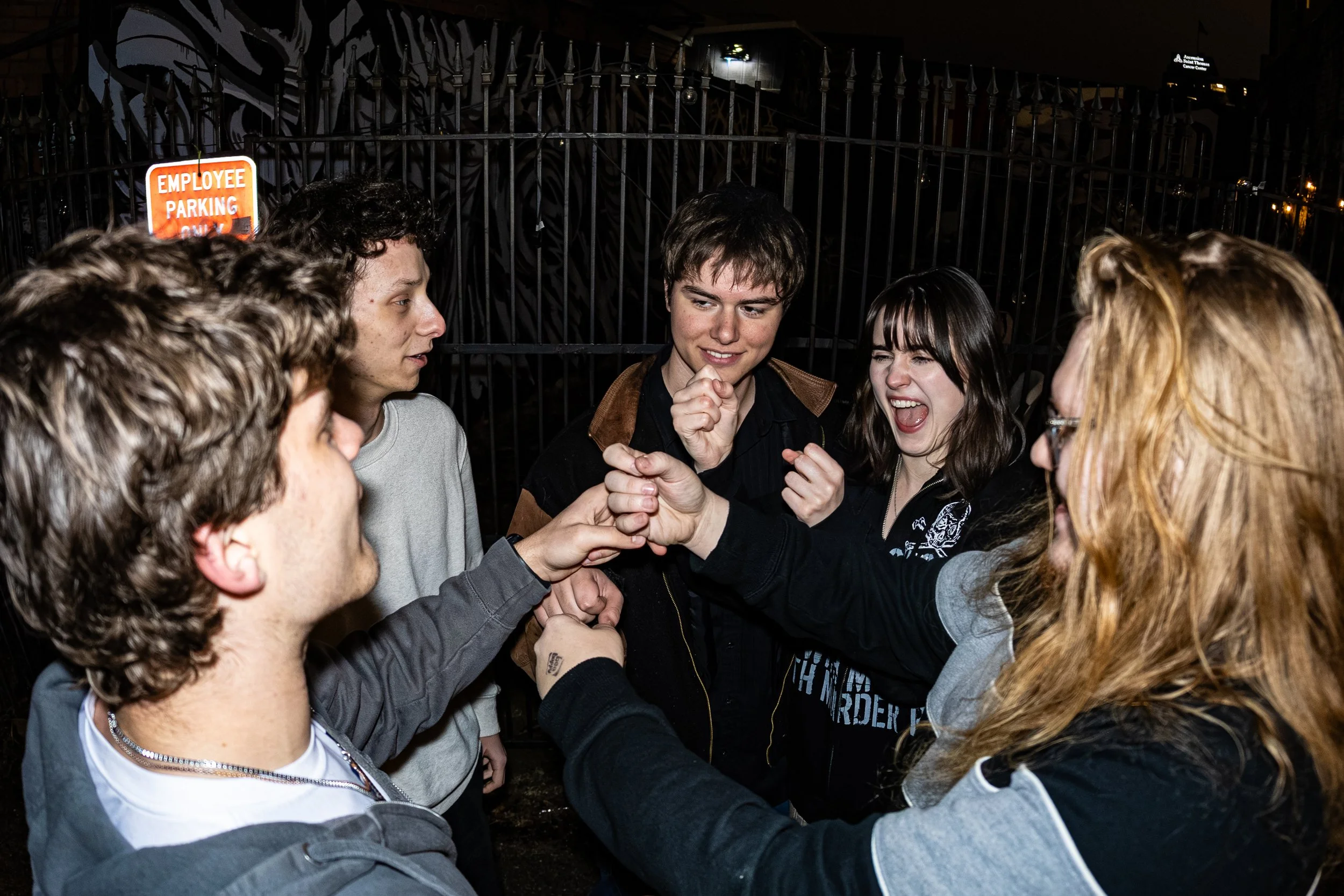 Group of young people playfully competing in a handshake game outdoors at night near a black metal fence with a sign that says 'Employee Parking Only'.