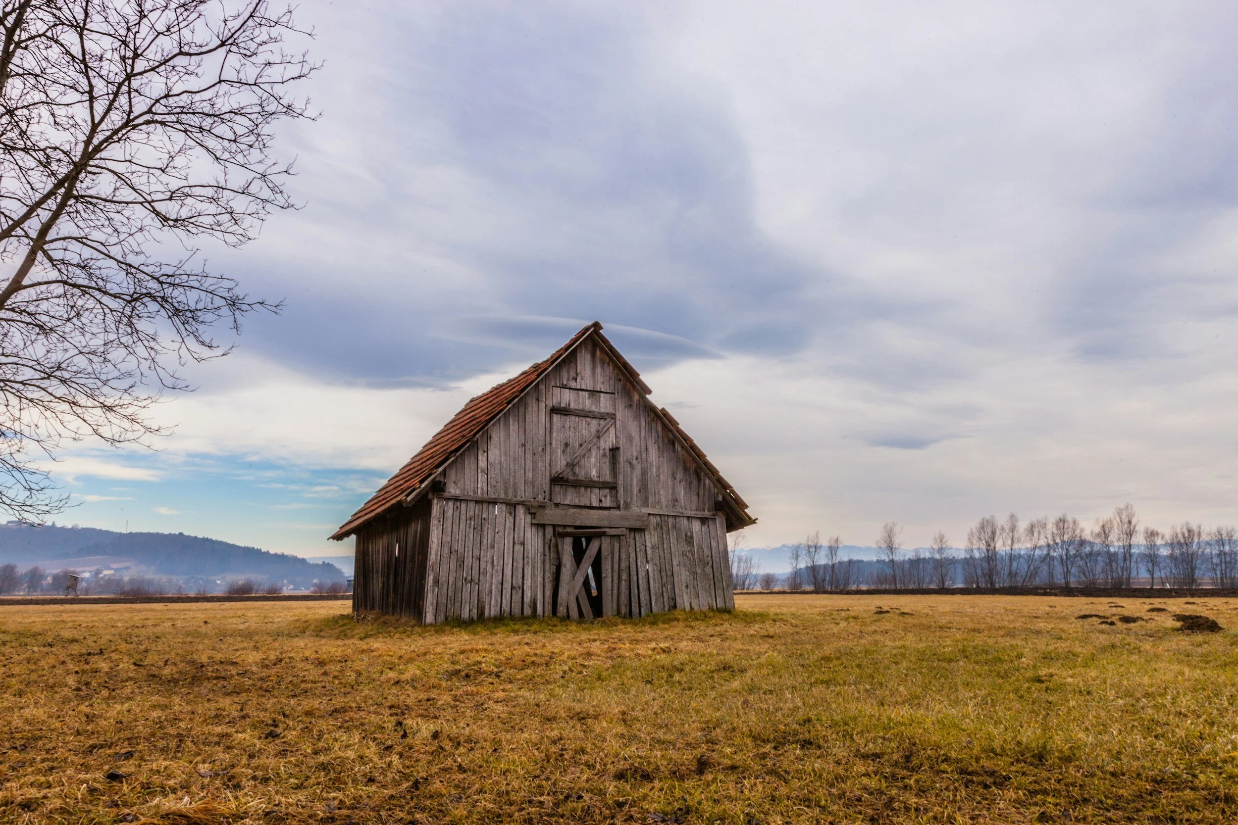 An old, weathered wooden barn with a crooked door in a vast open field under a cloudy sky.