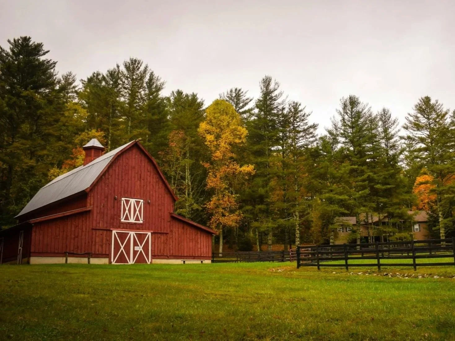 Red barn with white doors and trim, surrounded by green grass and trees with fall foliage under a cloudy sky.