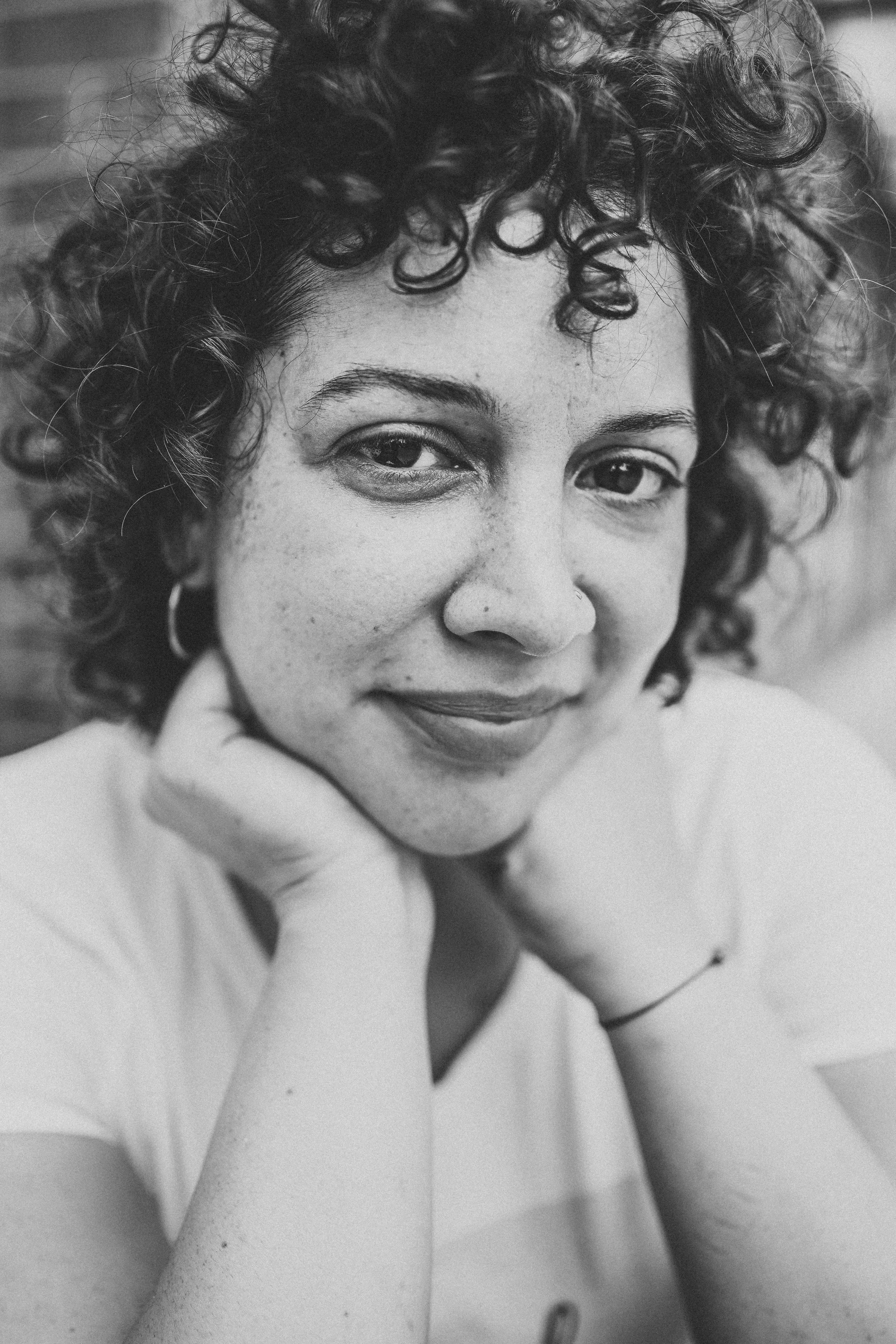 Black and white portrait of a woman with curly hair, smiling gently, resting her chin on her hands.