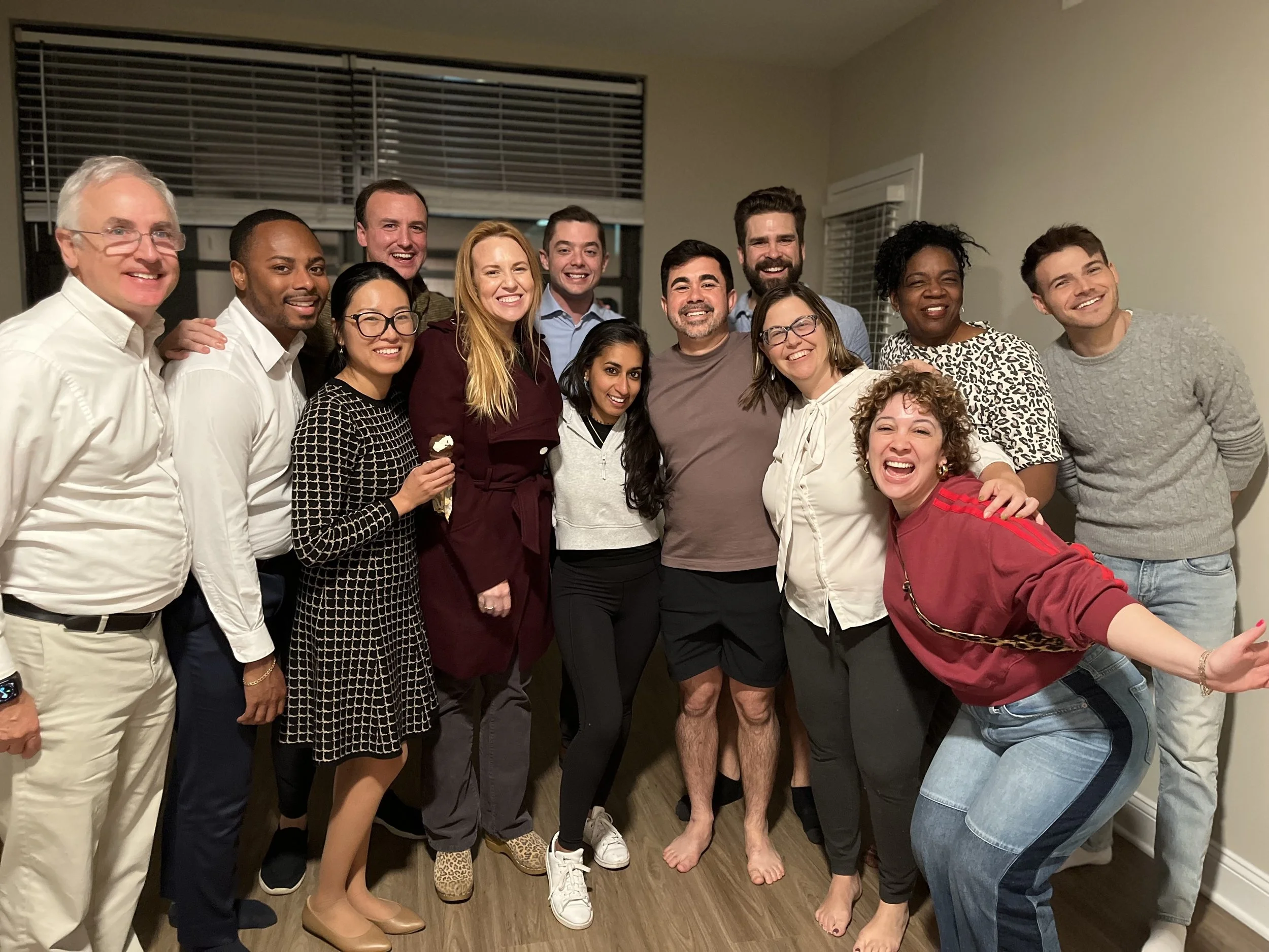 Group of 14 people standing together in a room, smiling and celebrating, with some making playful gestures.