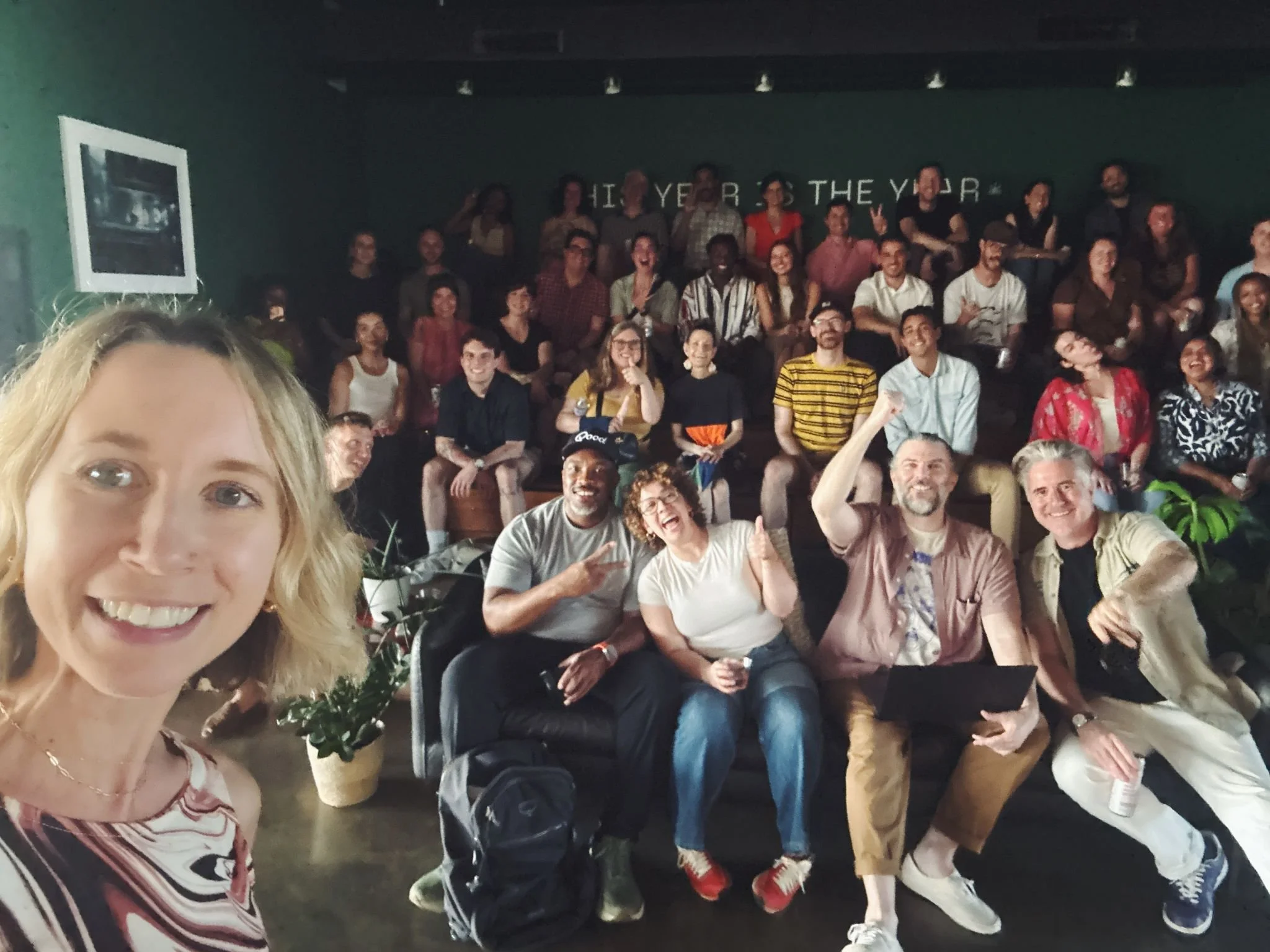 A group selfie of smiling people in a dimly lit indoor space, with some sitting on couches and others standing behind, posing for the camera.