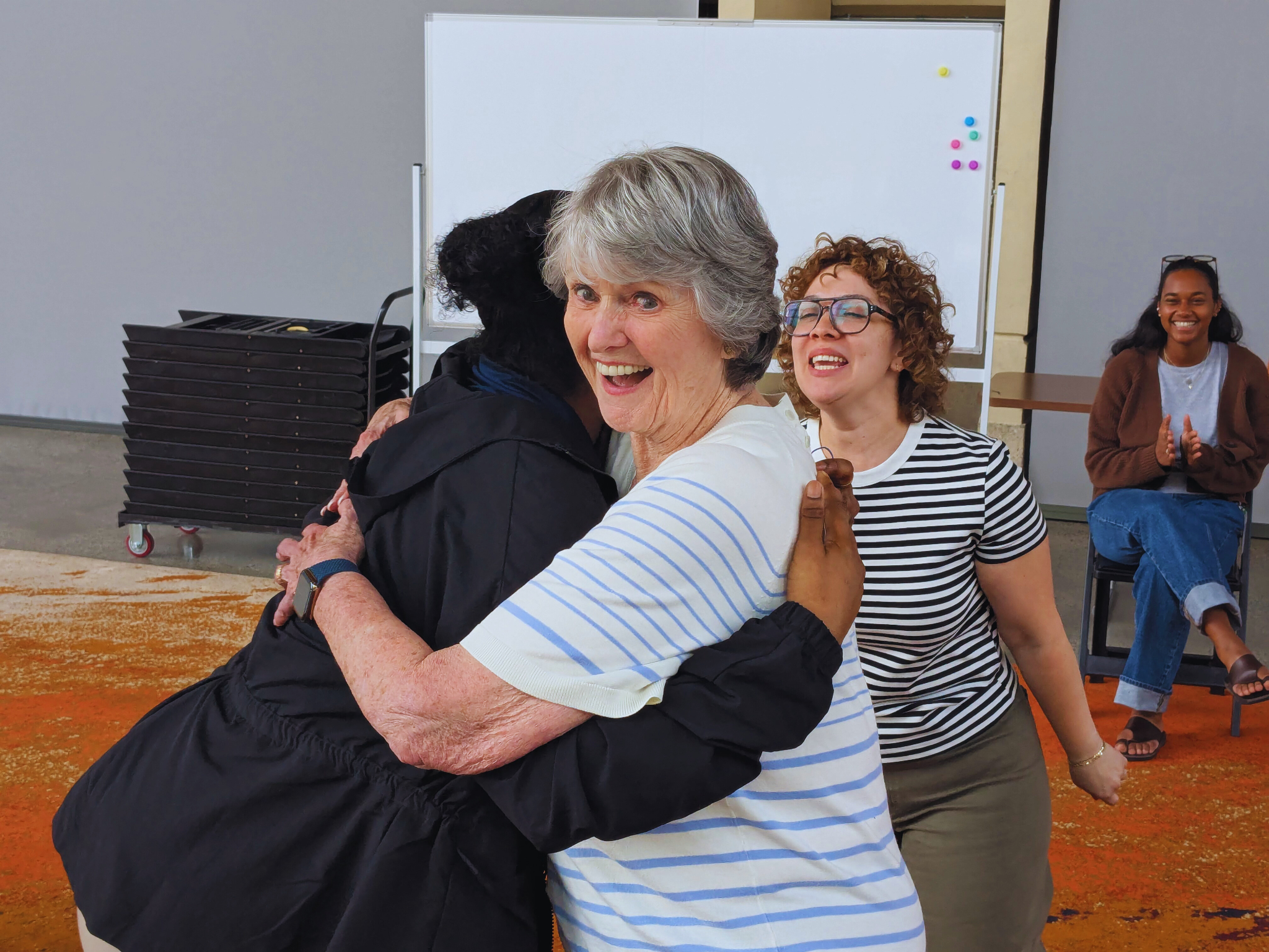 People hugging and smiling at a gathering with a whiteboard and chairs in the background.