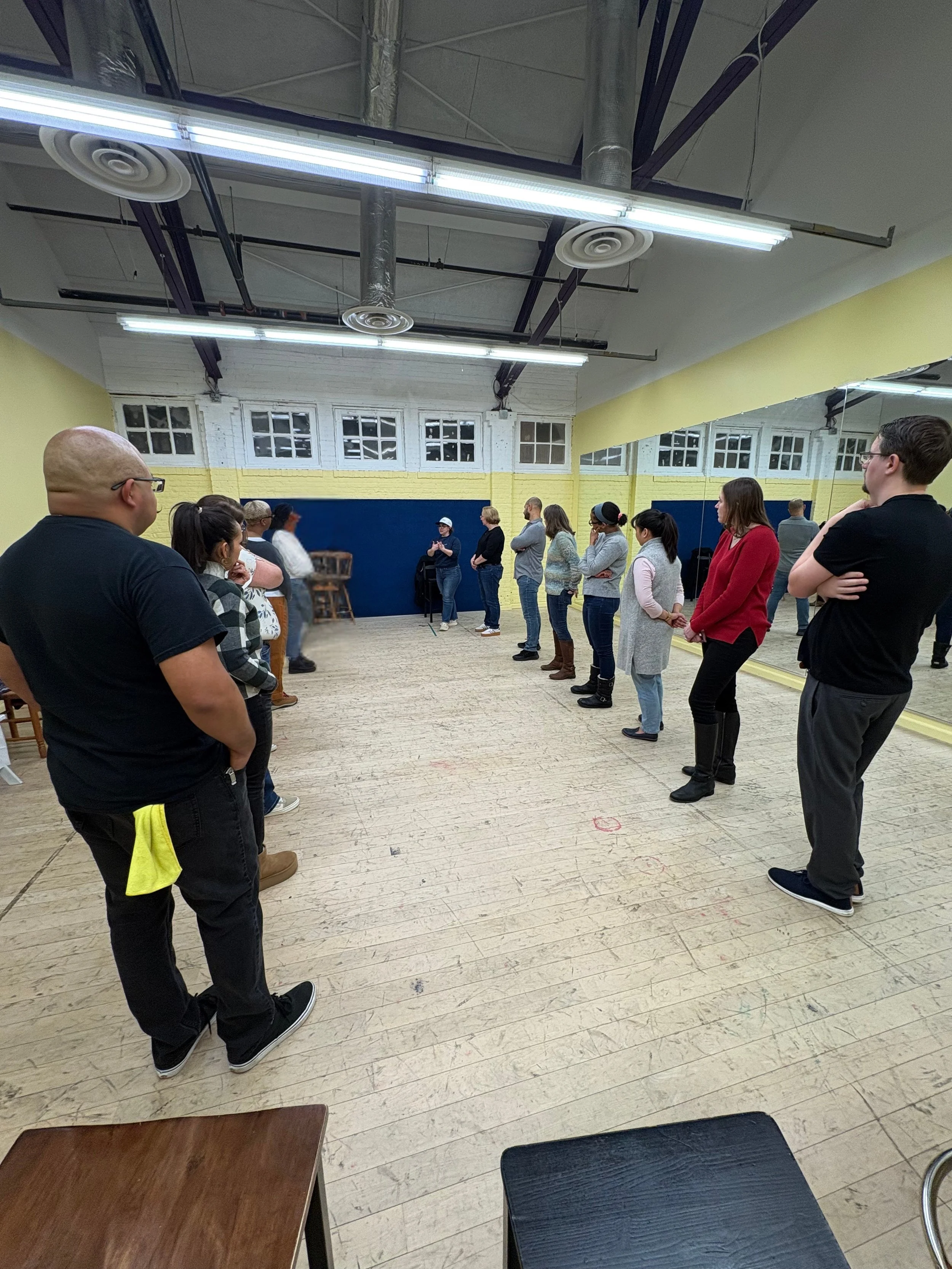 People standing in a spaced-out line in a dance or fitness studio with wood flooring and large wall mirrors.