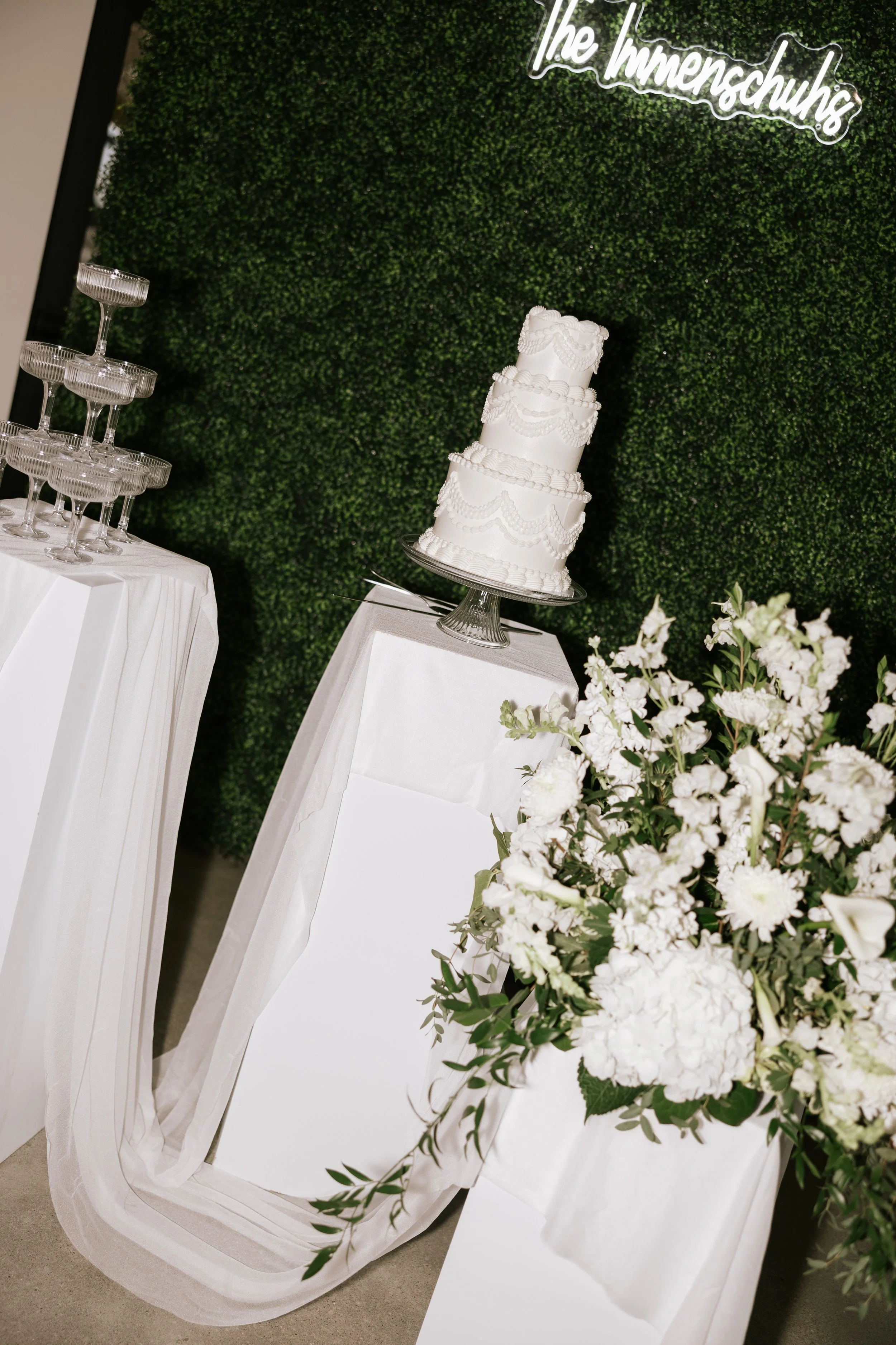A tiered white wedding cake with decorative icing, a bouquet of white flowers, a stack of empty glass dessert cups, and a green leafy backdrop with a neon sign reading 'The Innenschuhs'.