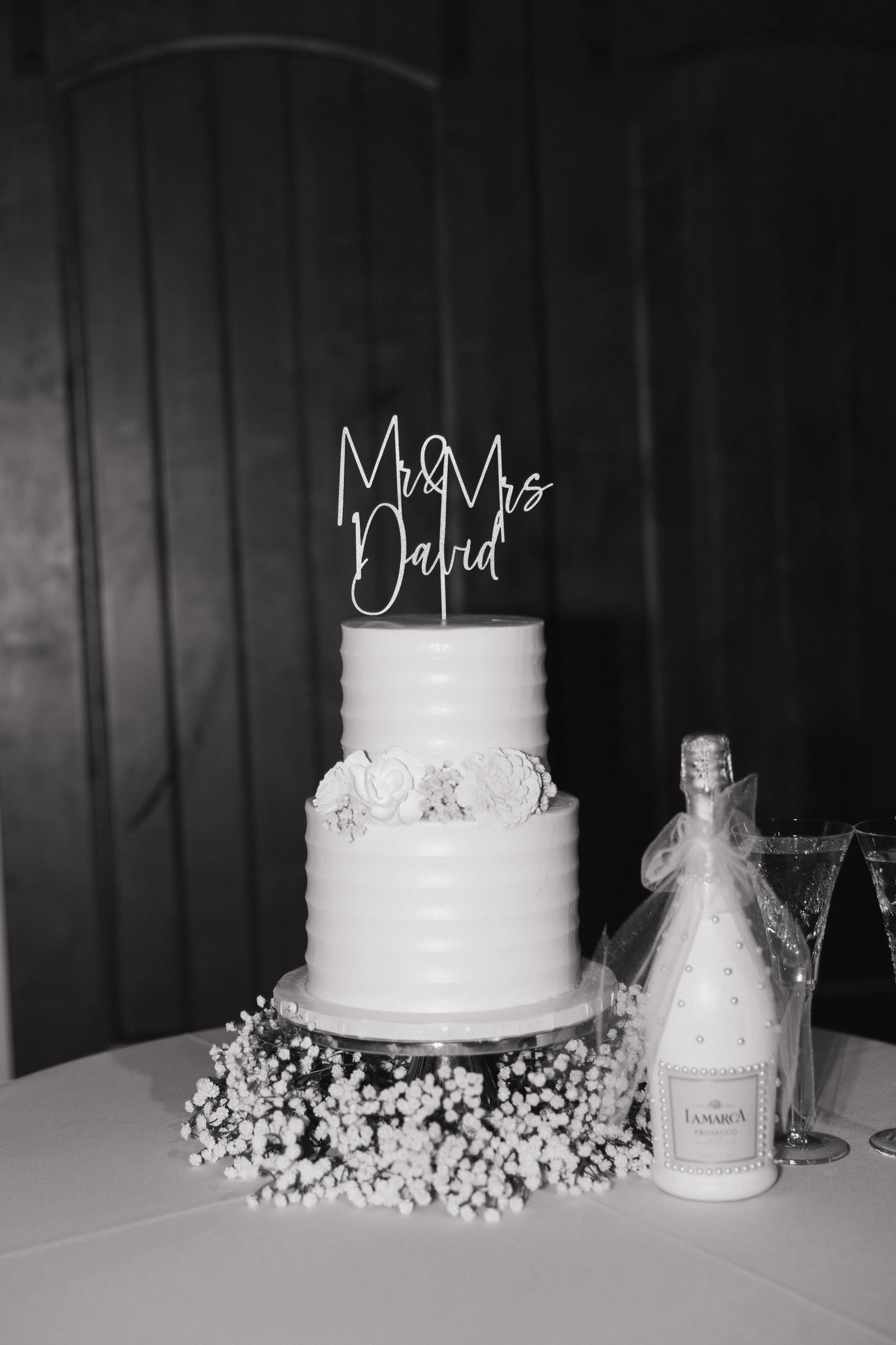 A two-tiered white wedding cake with floral decorations, topped with a cake topper reading 'Mr & Mrs Dalra,' surrounded by a ring of small white flowers, and a bottle of champagne with a wrapped ribbon on it.
