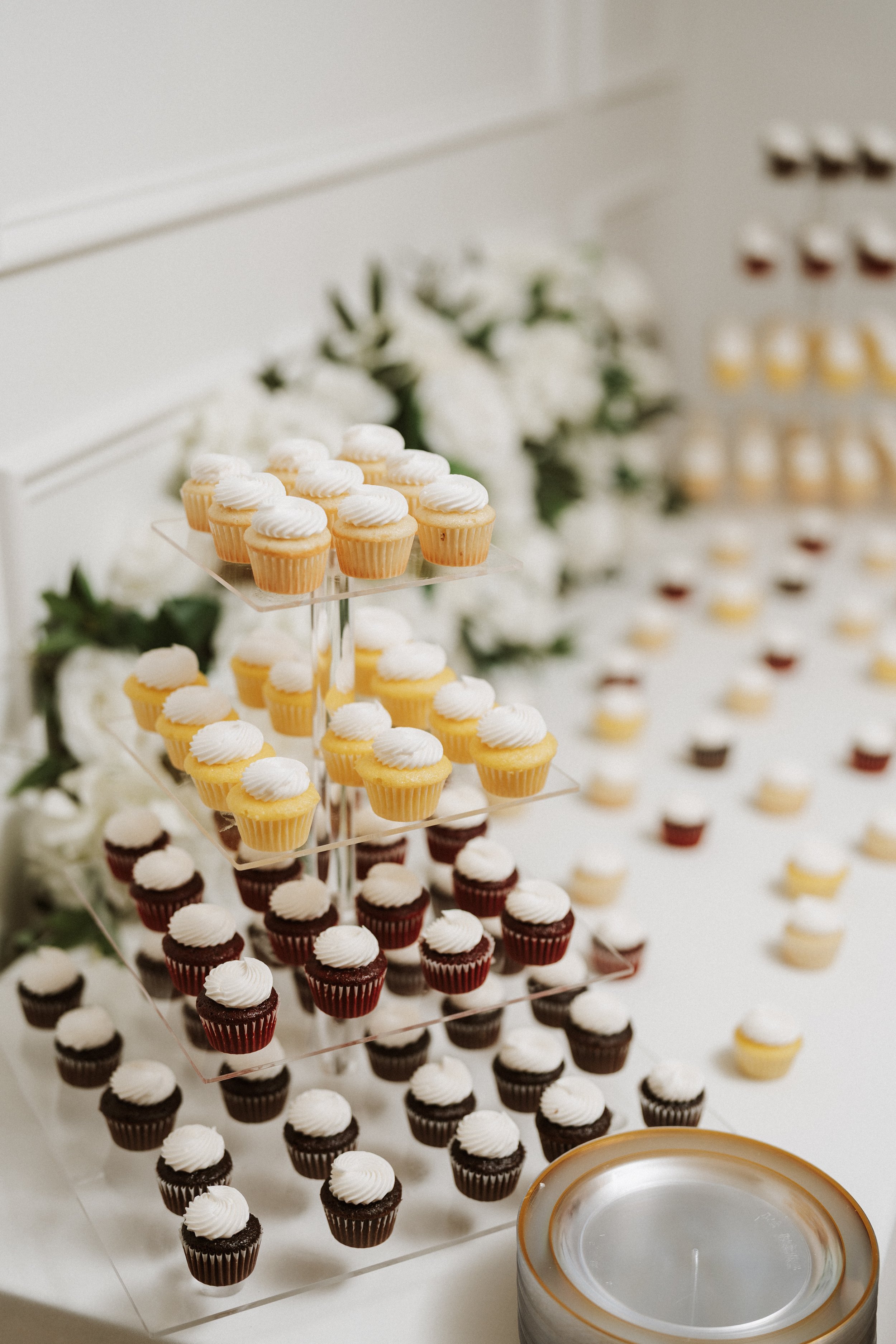 A display of cupcakes on a tiered stand with white frosting, arranged in three rows by flavor, in front of a white wall with floral decorations.