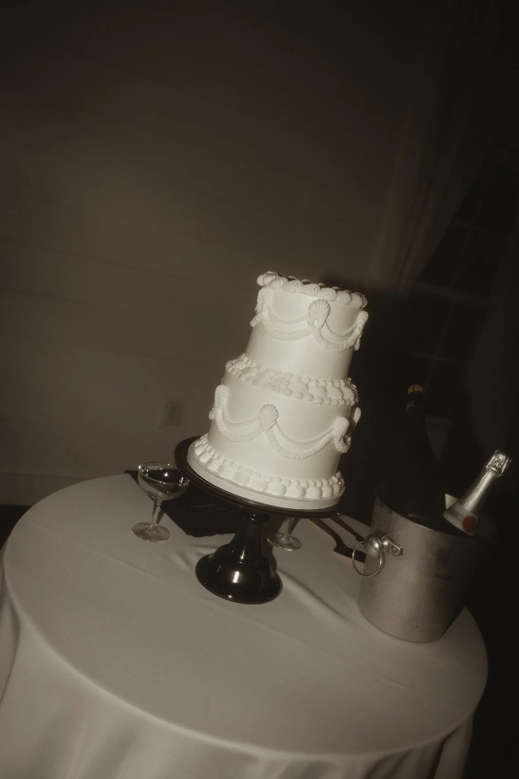 A three-tiered white wedding cake with decorative white piping, sitting on a black cake stand on a round white table, alongside a bottle of champagne in an ice bucket and two empty glasses.