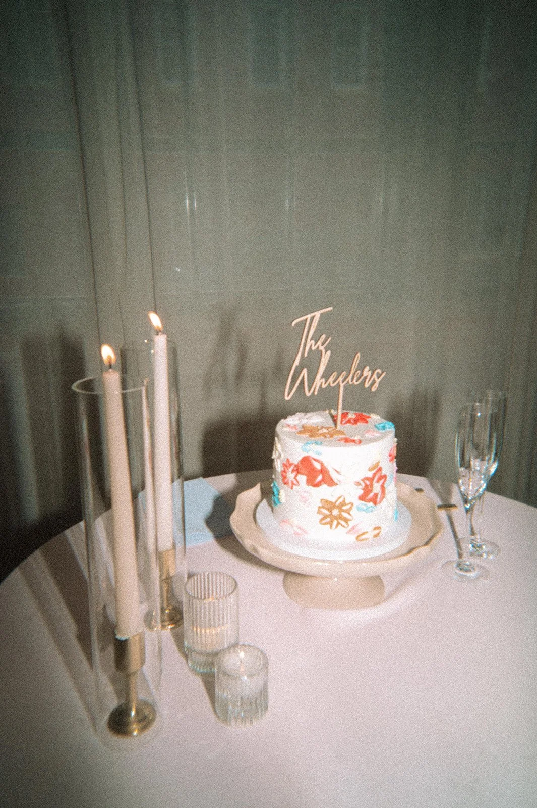 A white cake with colorful flower designs and a topper that says "The Wheelers" sits on a plate on a white tablecloth with candles and champagne glasses nearby.