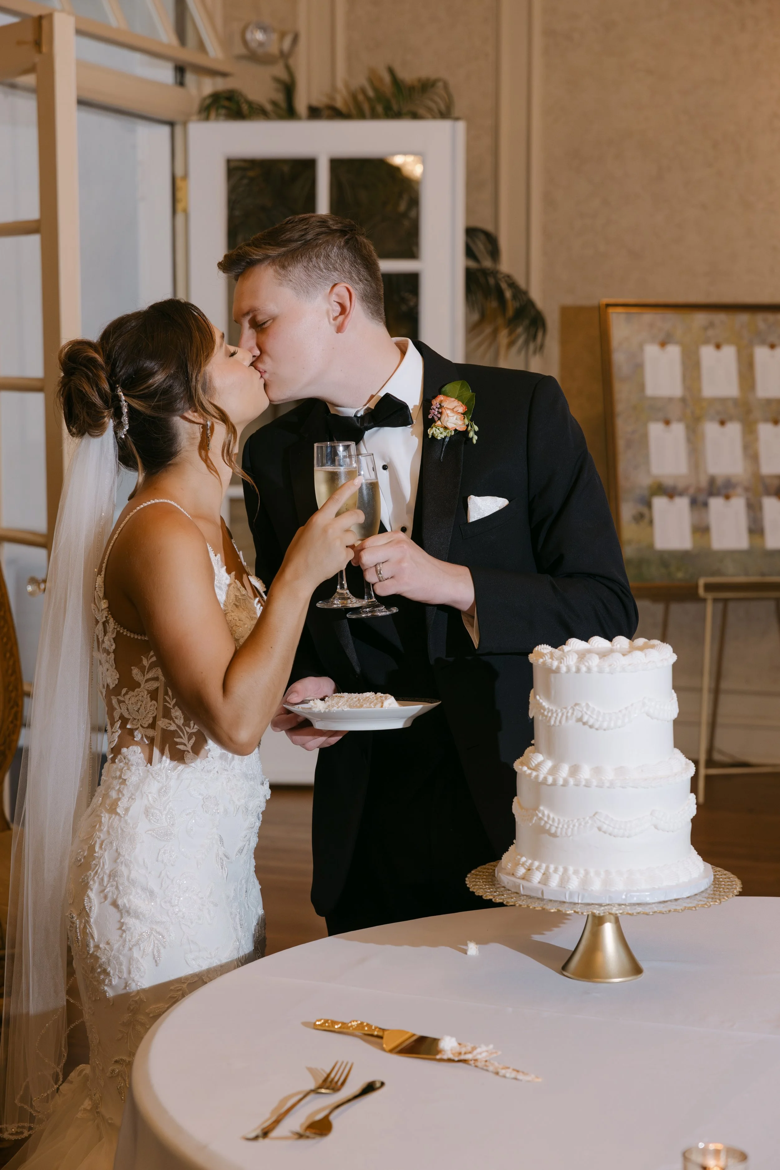 Newlywed bride and groom sharing a kiss during their wedding reception, holding champagne glasses, with a wedding cake on the table.