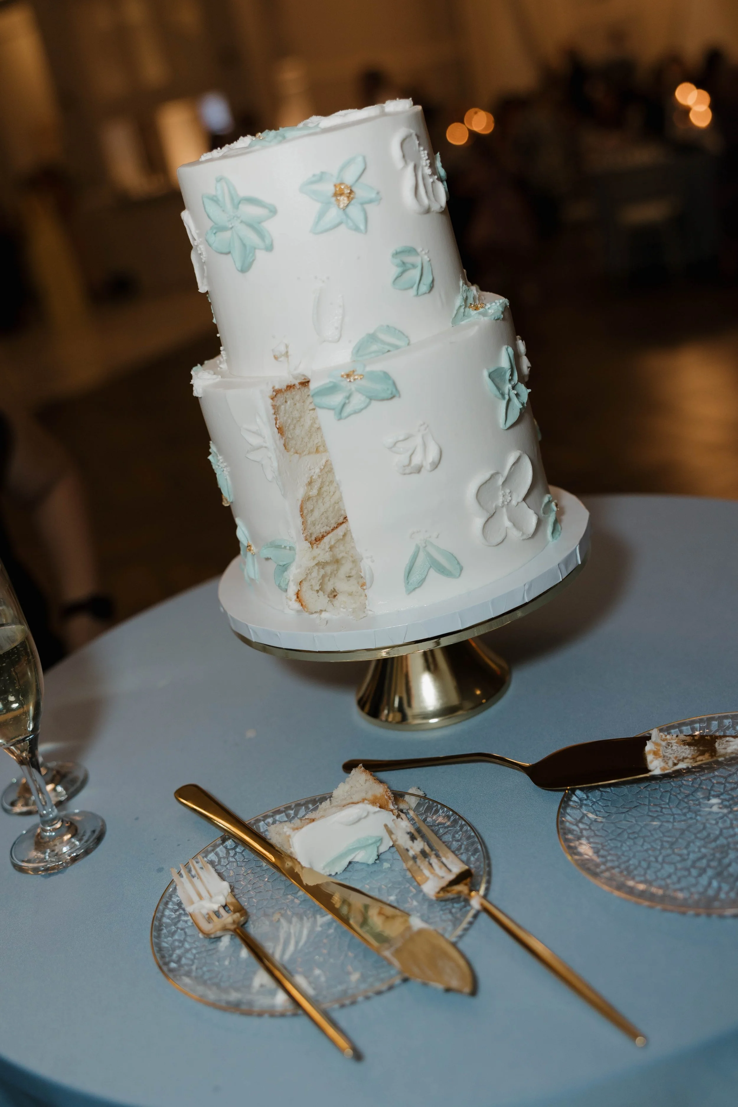 A tall, two-tiered white wedding cake decorated with light blue flowers and gold accents, with a slice removed showing the inside. The cake is on a gold cake stand on a blue tablecloth, with a slice cut and placed on a clear glass plate with gold ute