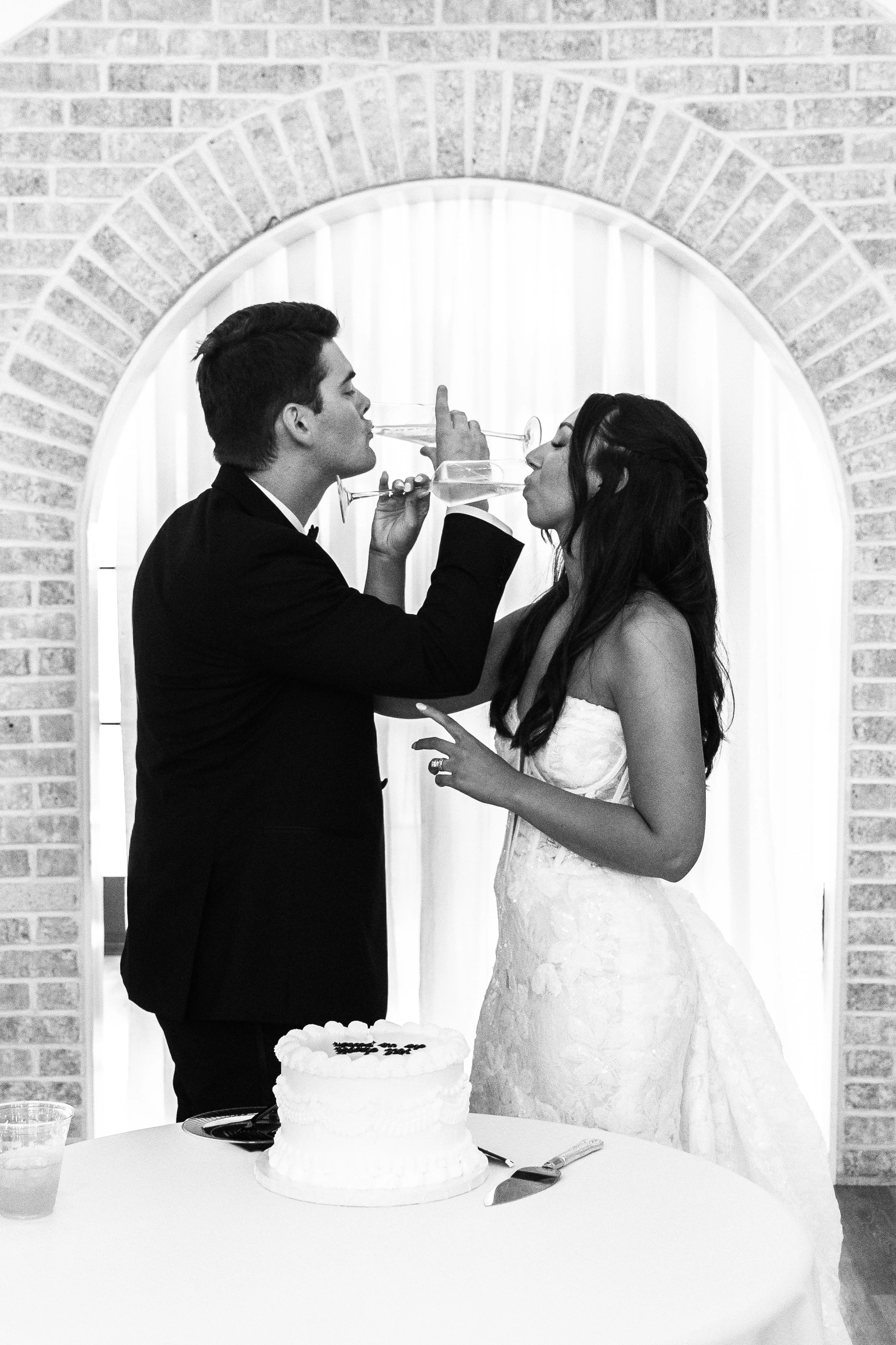 A black-and-white photo of a bride and groom at their wedding reception, holding champagne glasses and touching tips, standing in front of a curtain with an arched brick wall behind them. A wedding cake with a knife and a glass of drink are on the ta